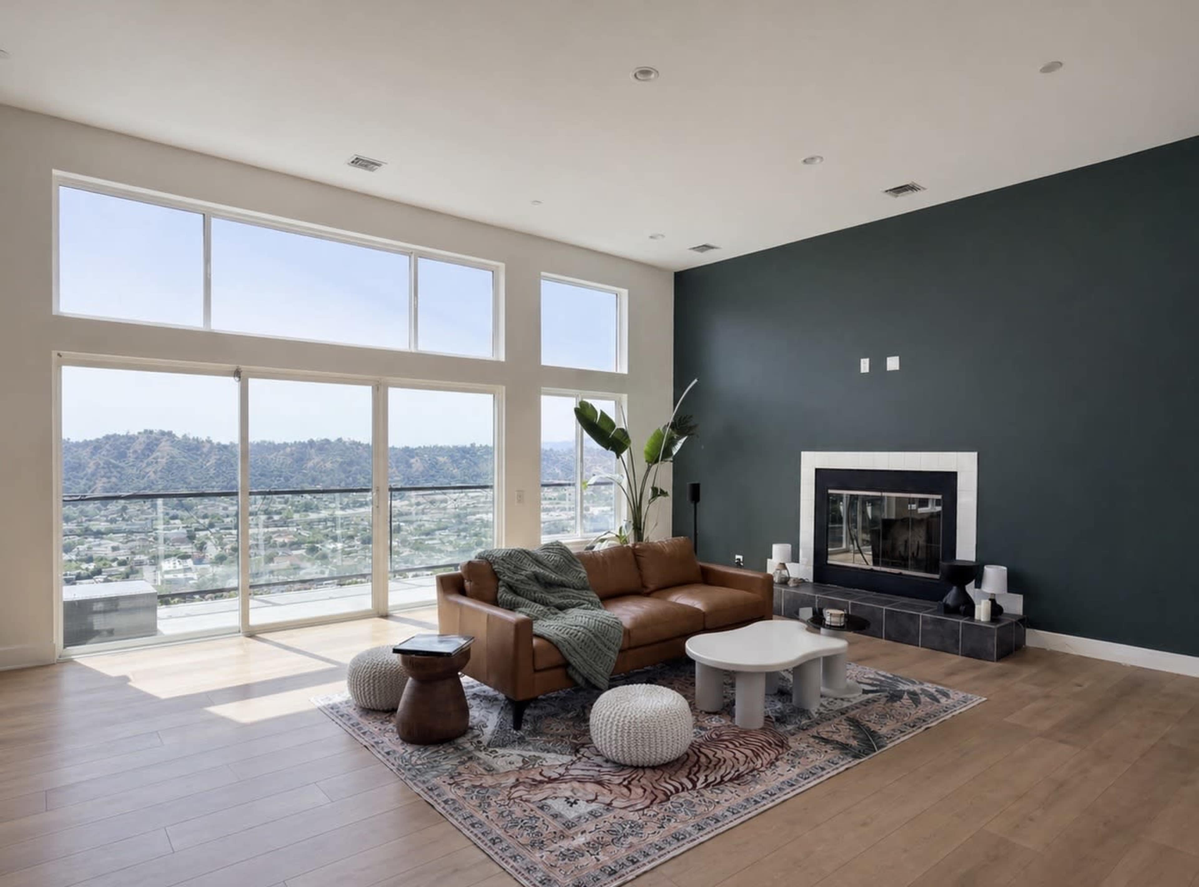 The image shows a modern living room with a brown leather sofa, a white coffee table, a fireplace, and large windows overlooking a mountain view.