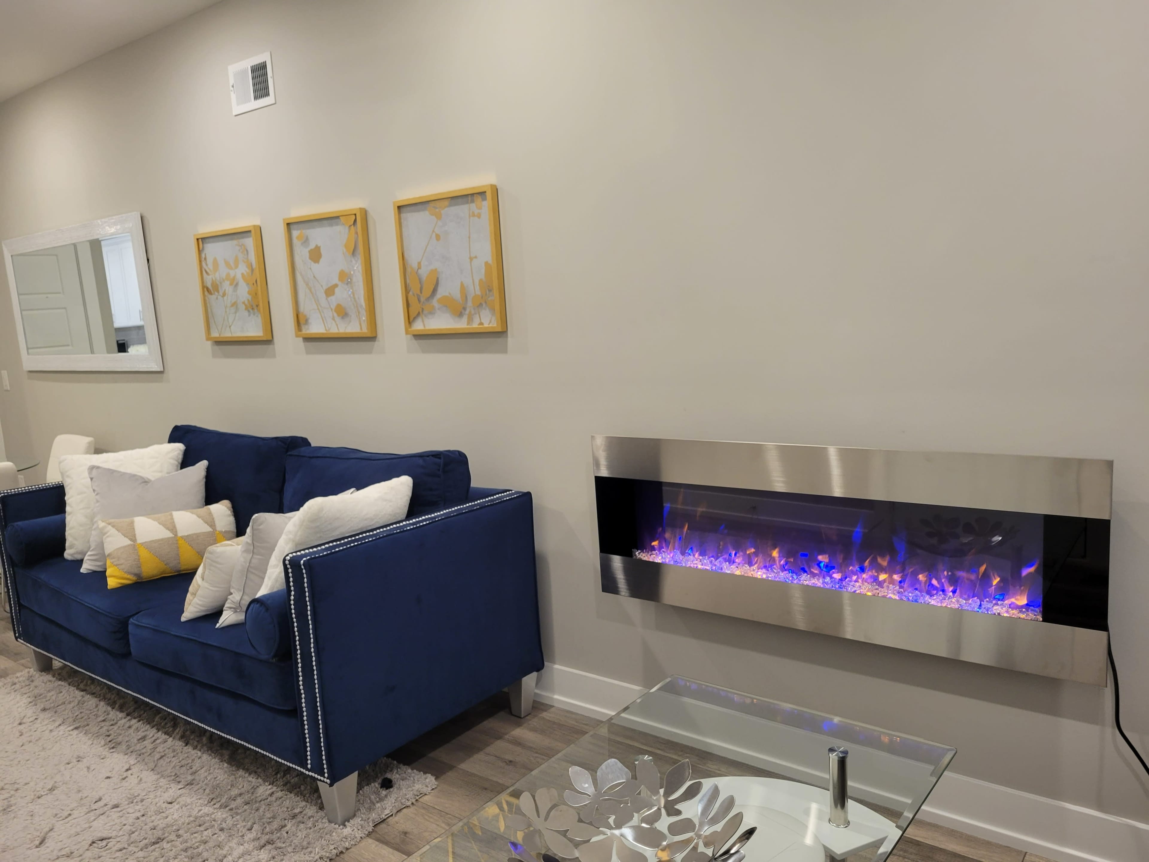 A blue sofa with white and yellow decorative pillows is positioned next to a modern wall-mounted electric fireplace, with framed artworks hanging above it on a light-colored wall.