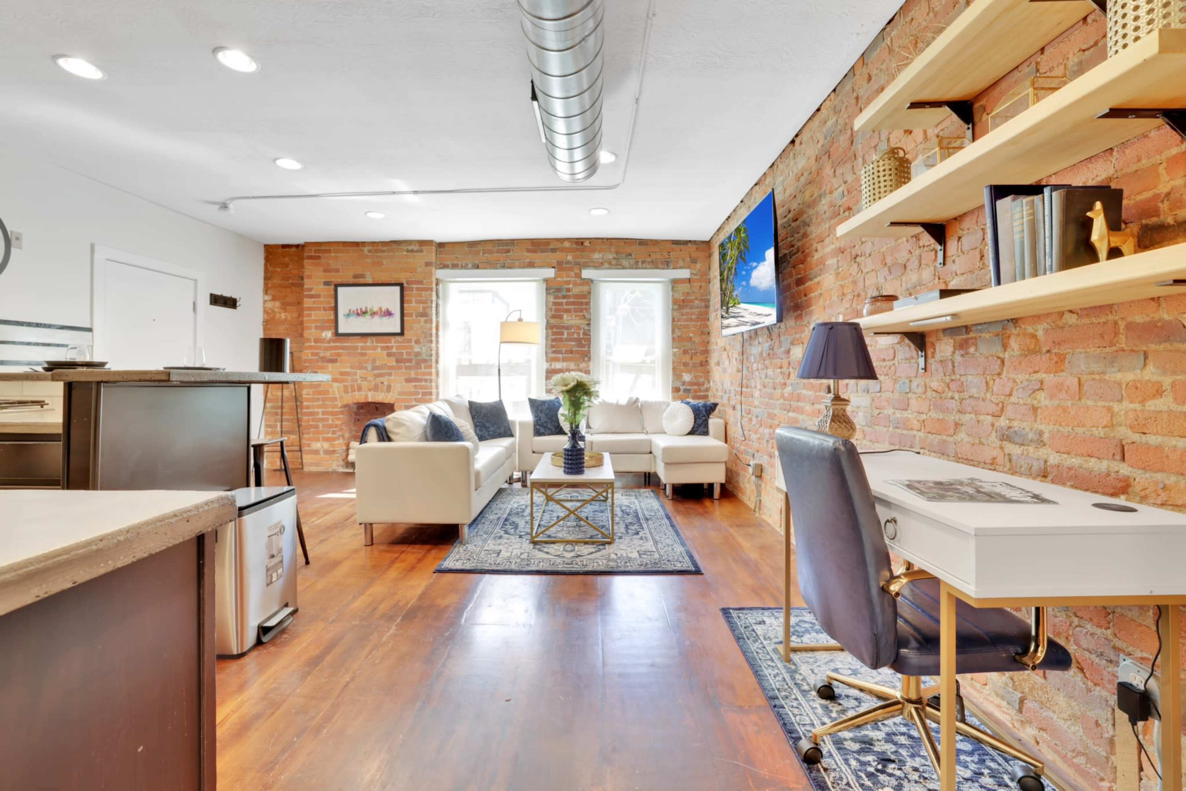 A bright living area with exposed brick walls, a sofa, a desk, and a decorative shelf, all arranged on hardwood flooring.