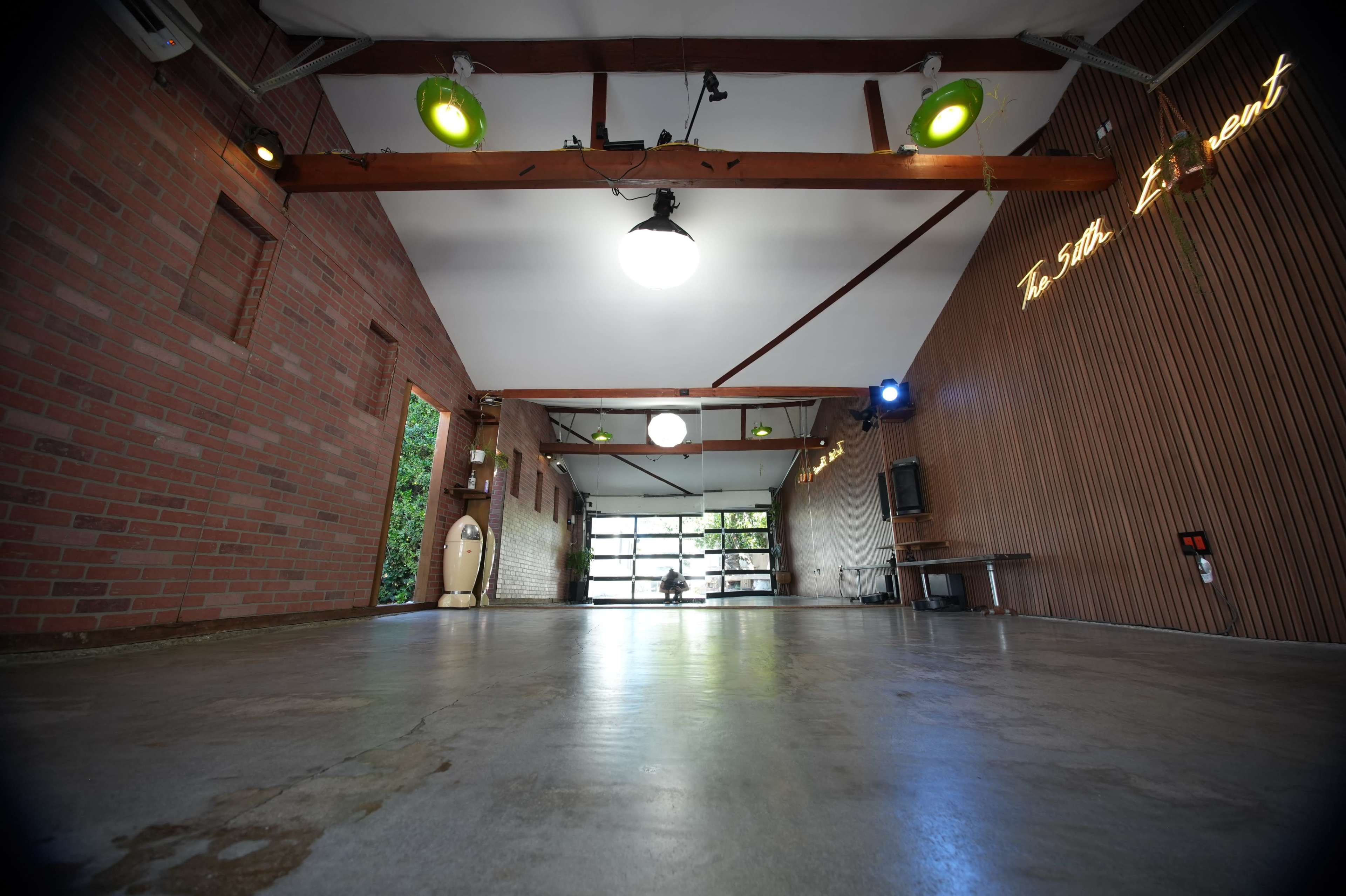 The image shows a spacious interior with brick walls, wooden beams, and a polished concrete floor, featuring overhead lighting and a large garage-style door.