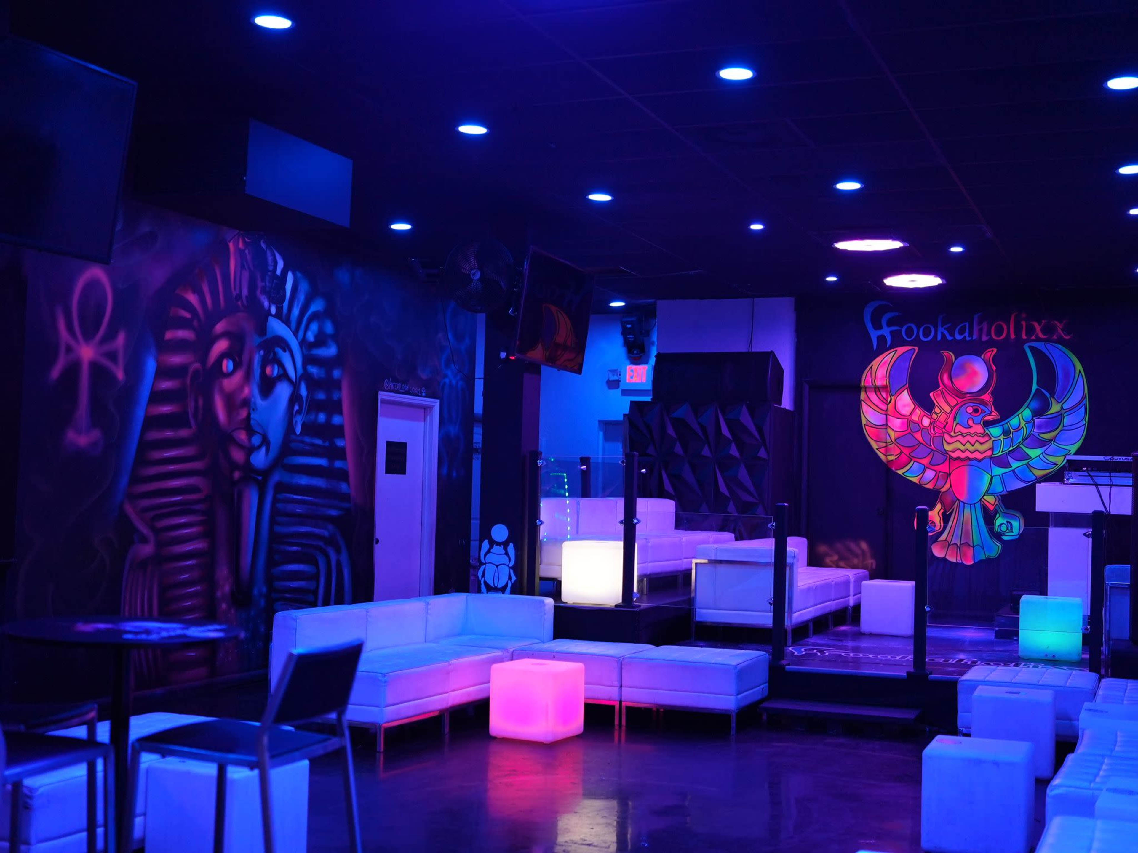 A dimly lit lounge with white seating, colorful lights, and vibrant mural artwork on the walls.