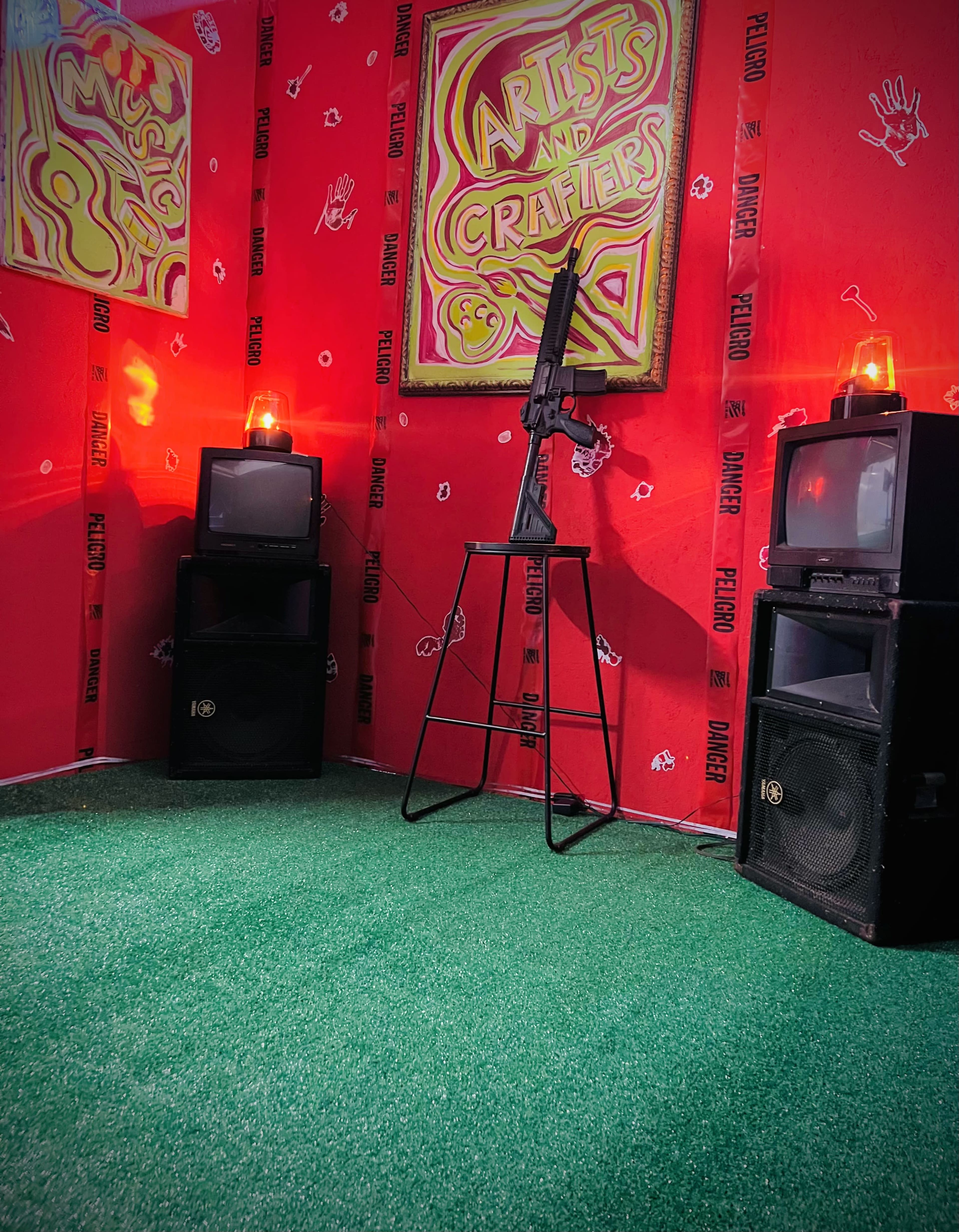 The image shows a corner of a room with red walls decorated with artwork that reads "Artists and Crafters," featuring a microphone stand and a rifle mounted on it, flanked by old televisions and speakers on either side.