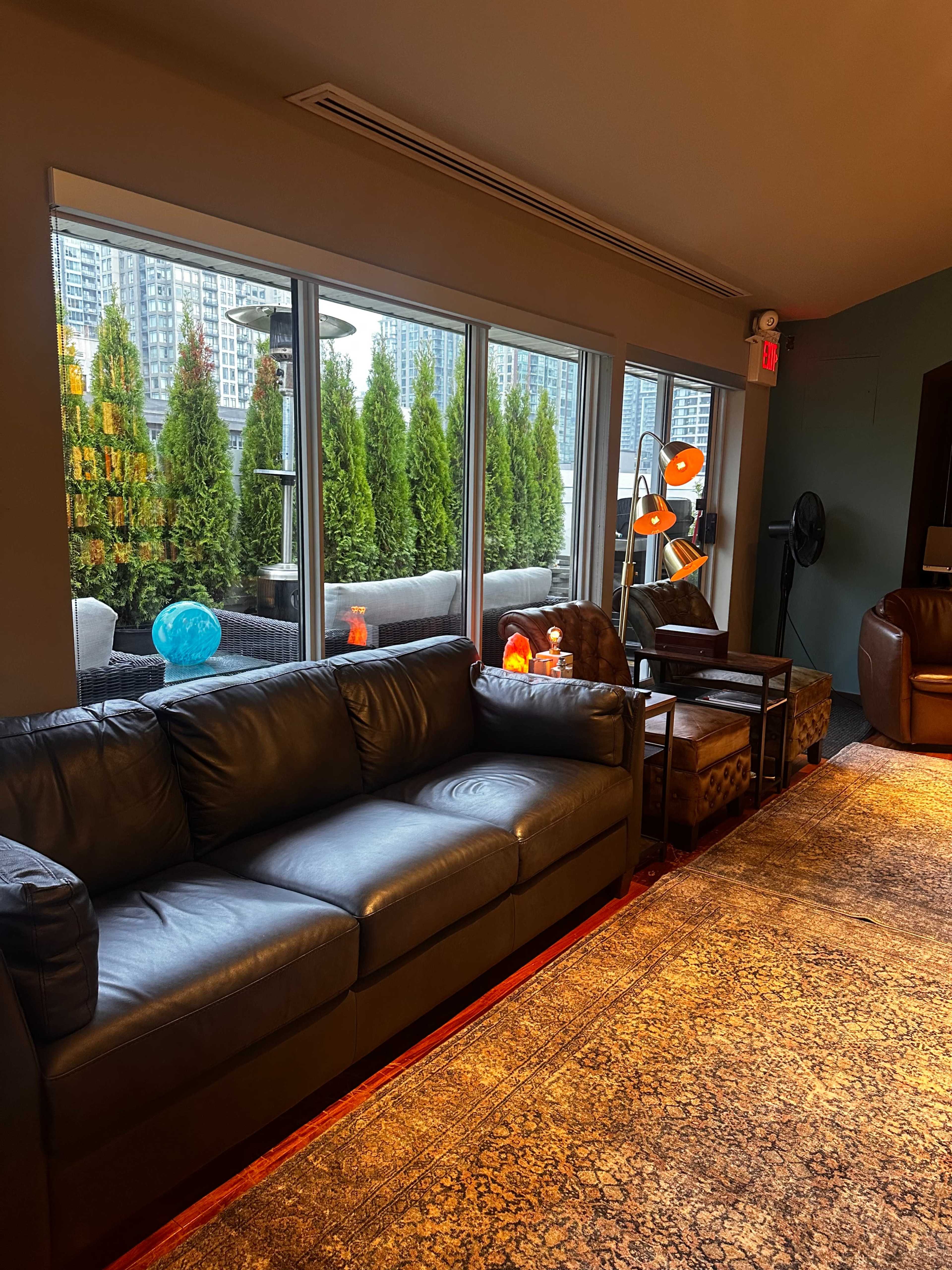 A black leather sofa is positioned in a cozy indoor seating area with large windows overlooking a landscaped outdoor space.