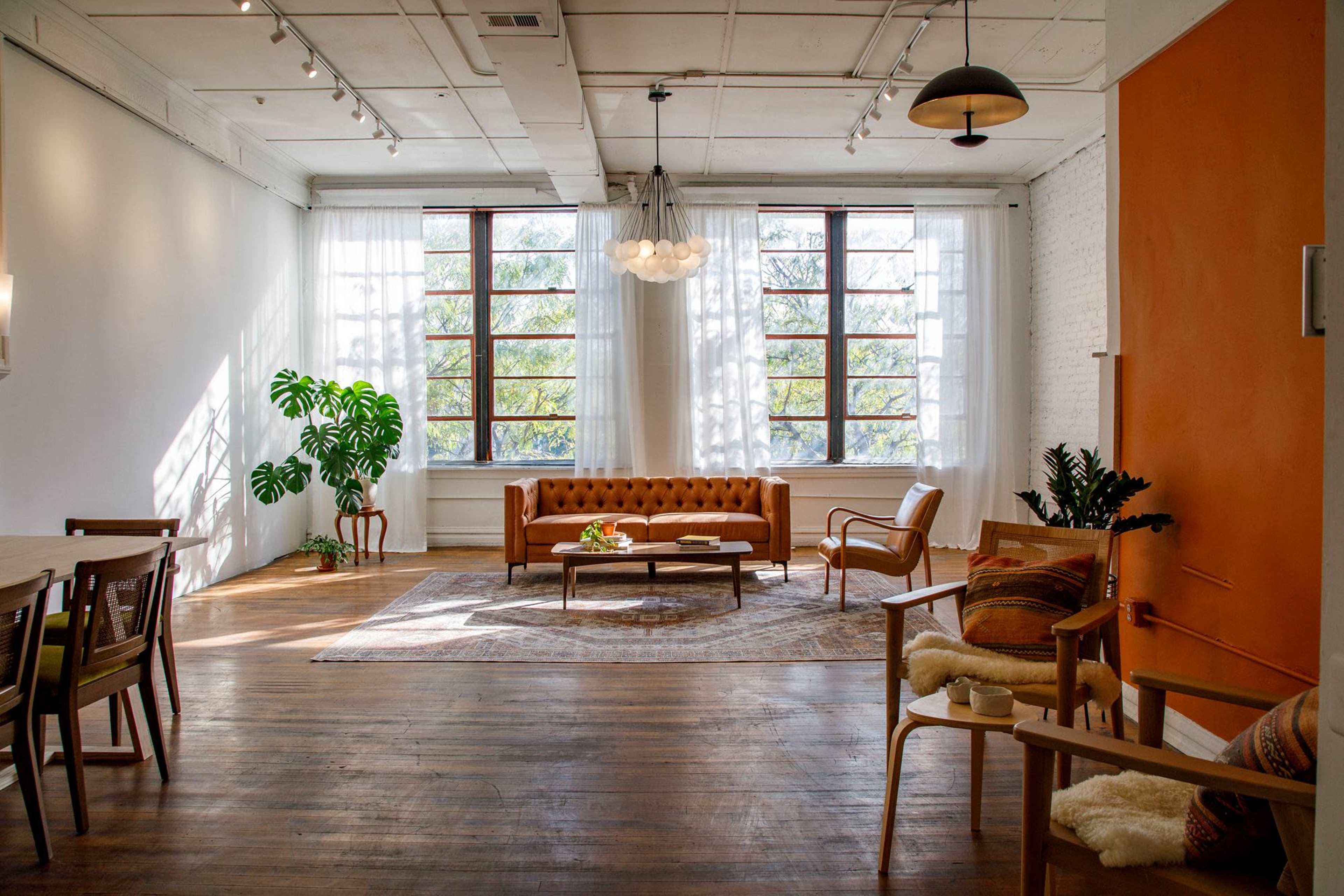 A spacious, well-lit room featuring a brown leather sofa, decorative chairs, a potted plant, and large windows with flowing curtains.