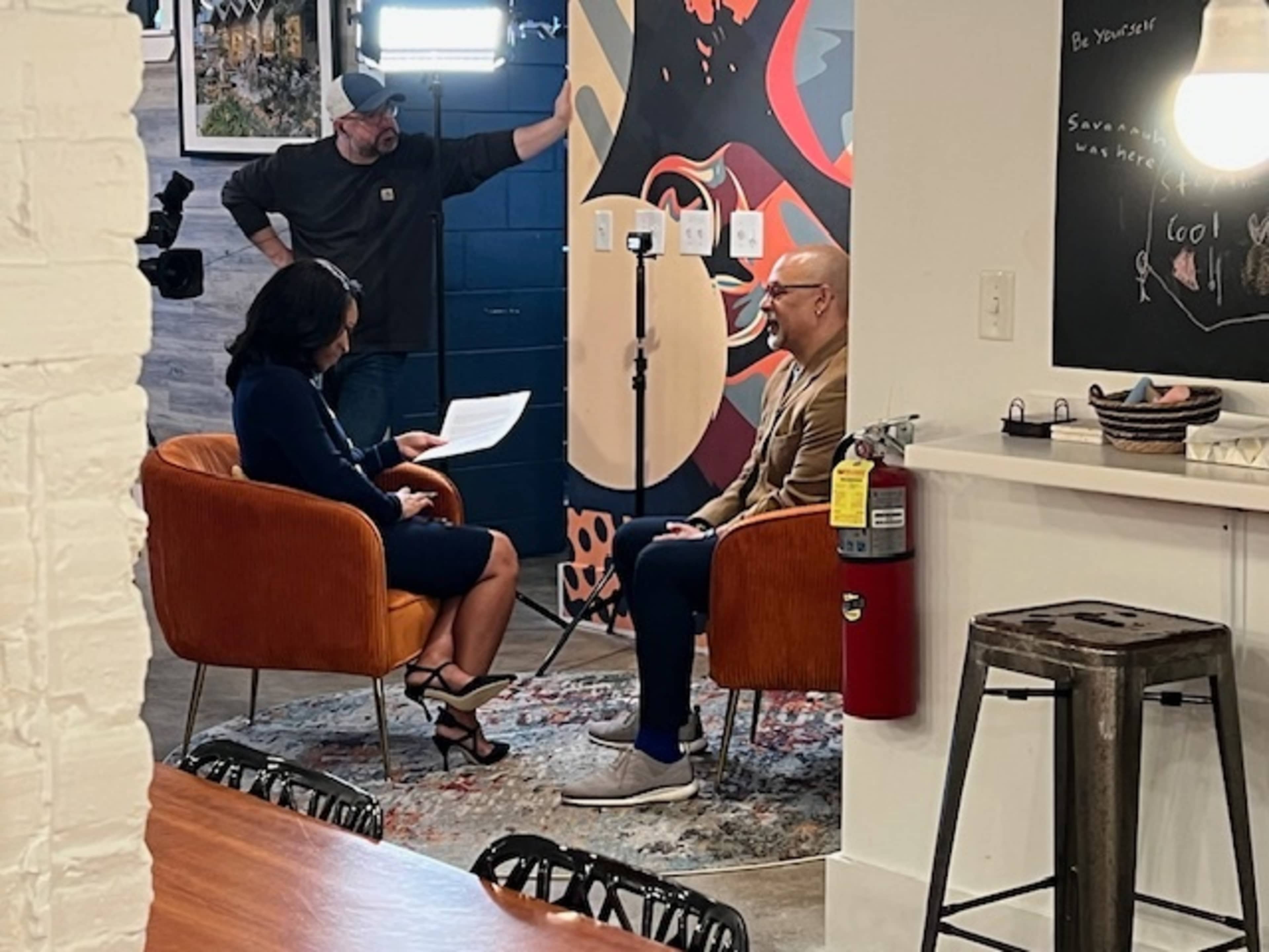 A woman in a dark dress interviews a man in a light-colored shirt while seated in modern orange chairs in a well-lit interior space.