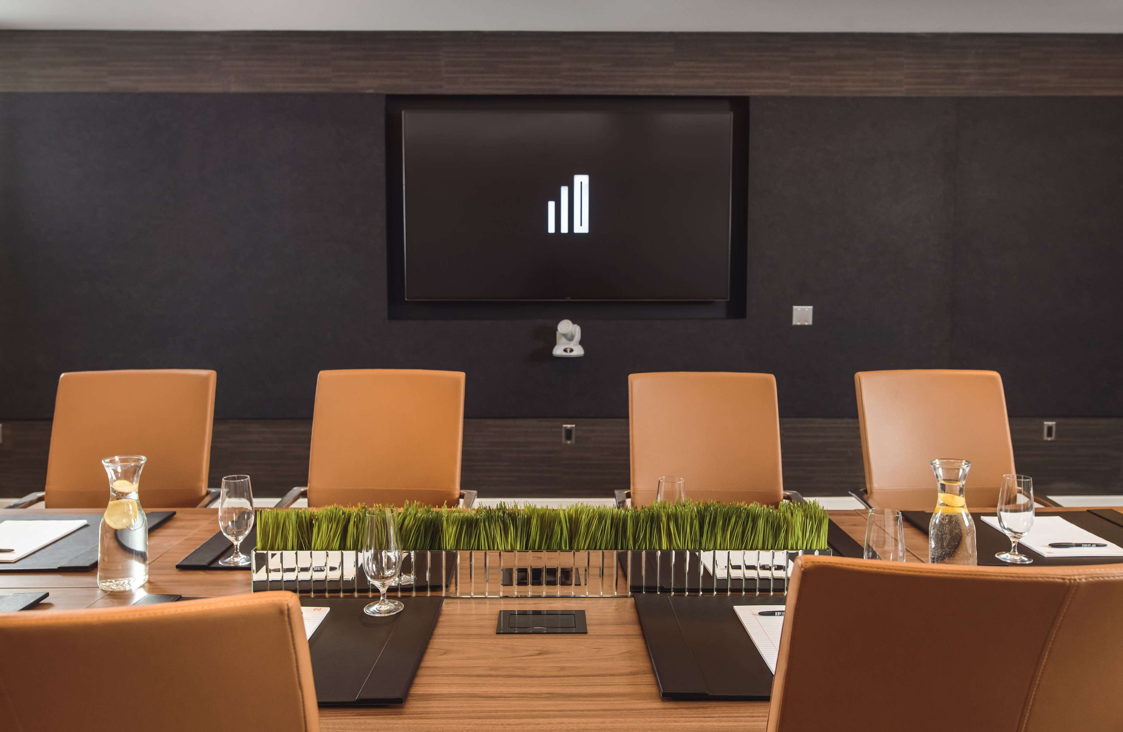 A modern conference room features a wooden table with orange chairs, a glass water pitcher, and a display screen showing a minimalist logo.