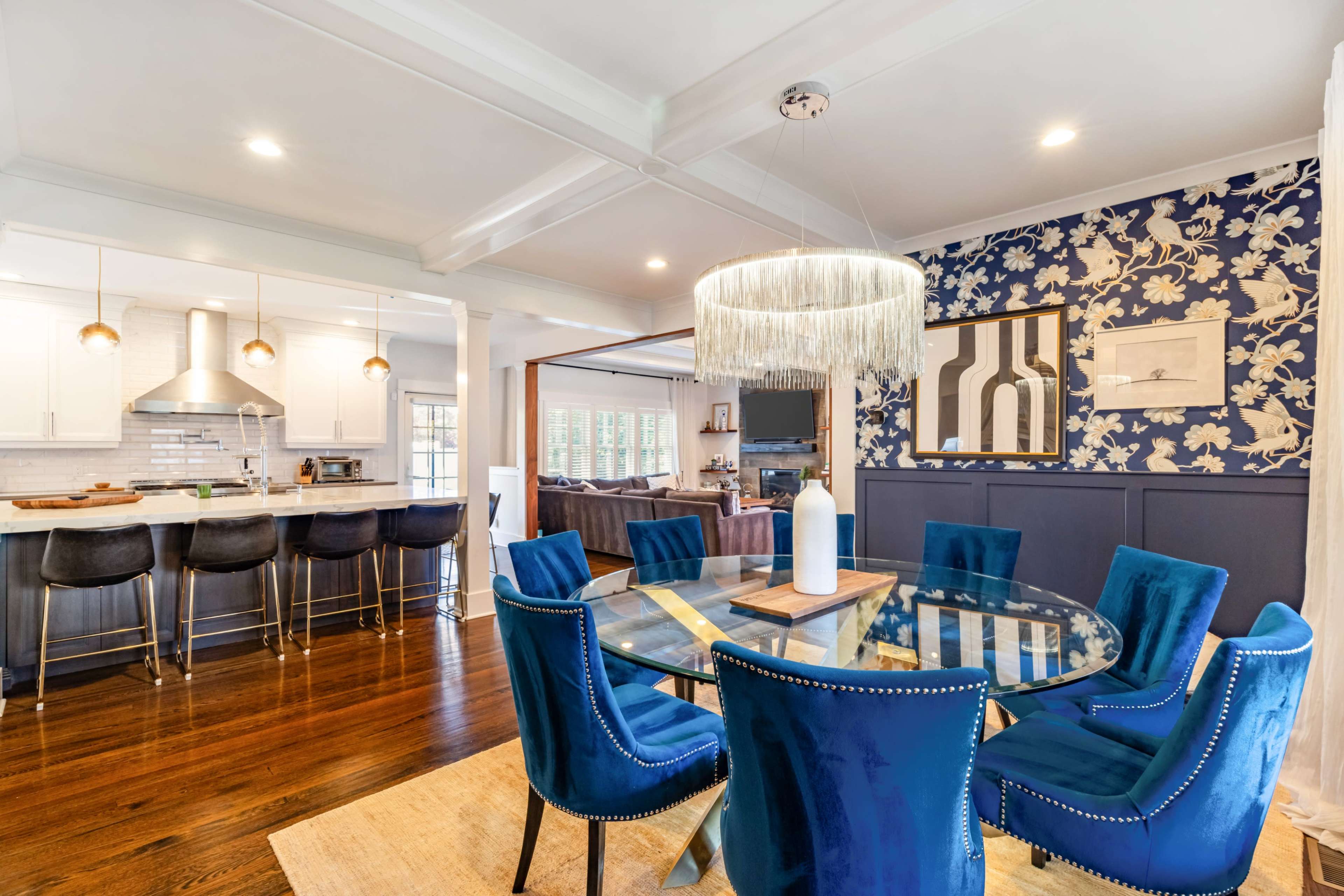 A modern dining area features a circular glass table surrounded by blue velvet chairs, with a patterned accent wall and a nearby kitchen visible in the background.