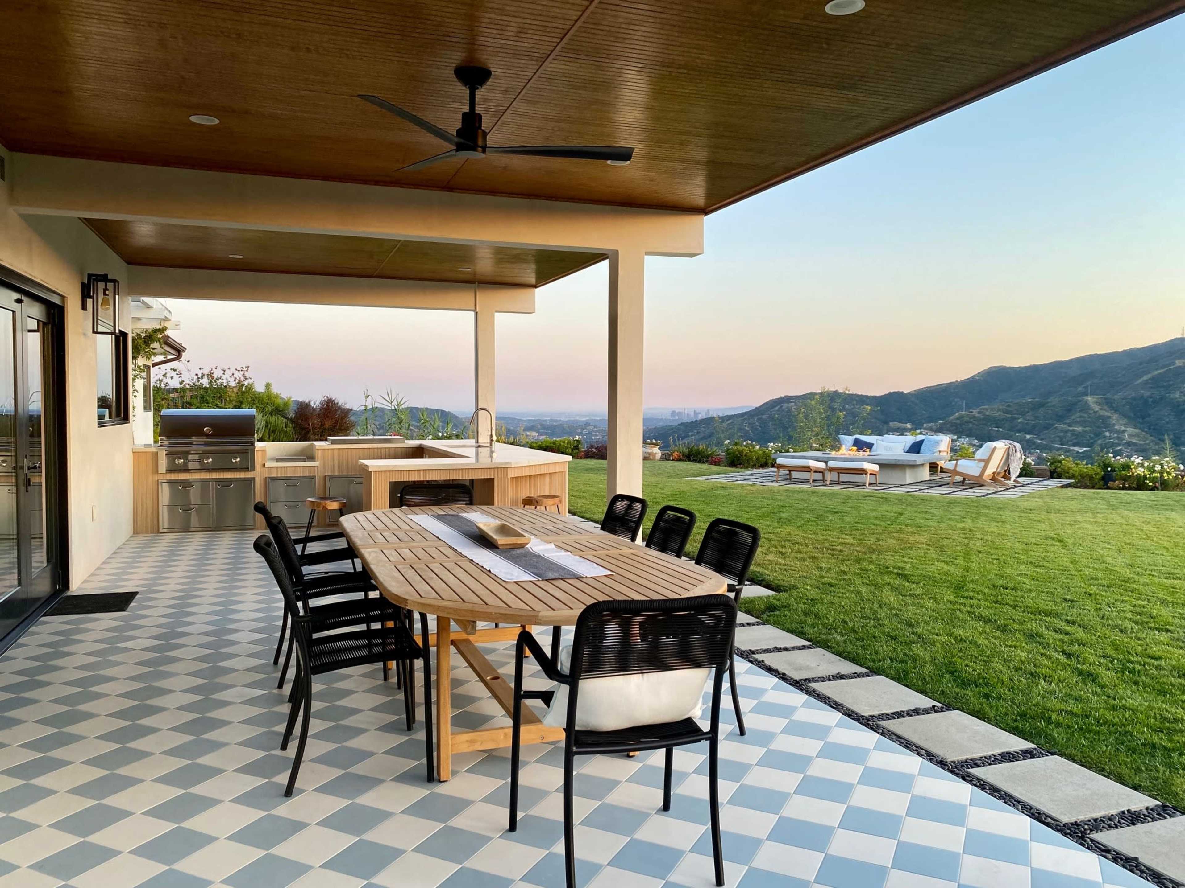 A patio features a large wooden dining table surrounded by black chairs, overlooking a lush green landscape and mountains under a clear sky.