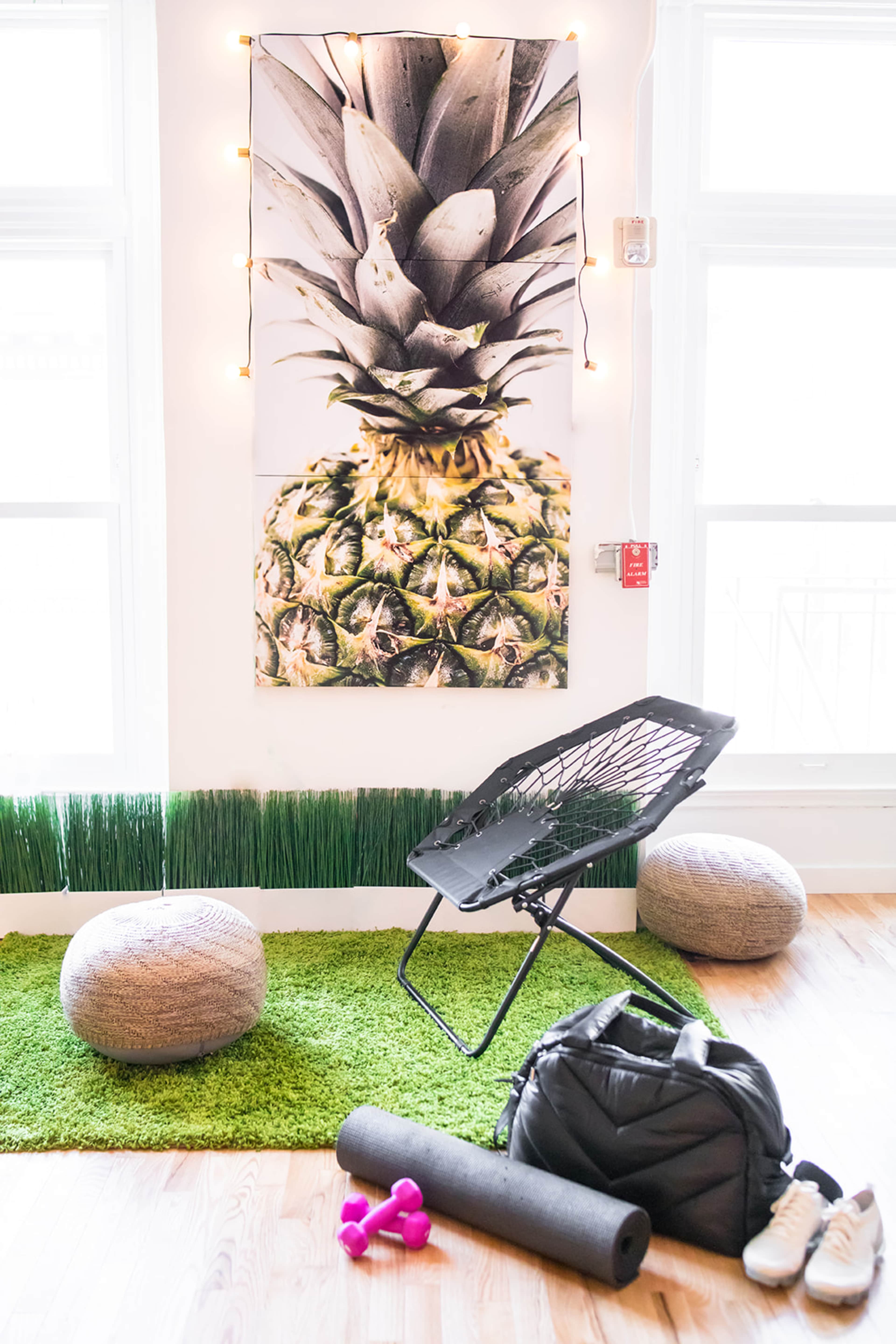 A room with a green rug, a chair, exercise equipment, and a large pineapple poster illuminated by string lights.