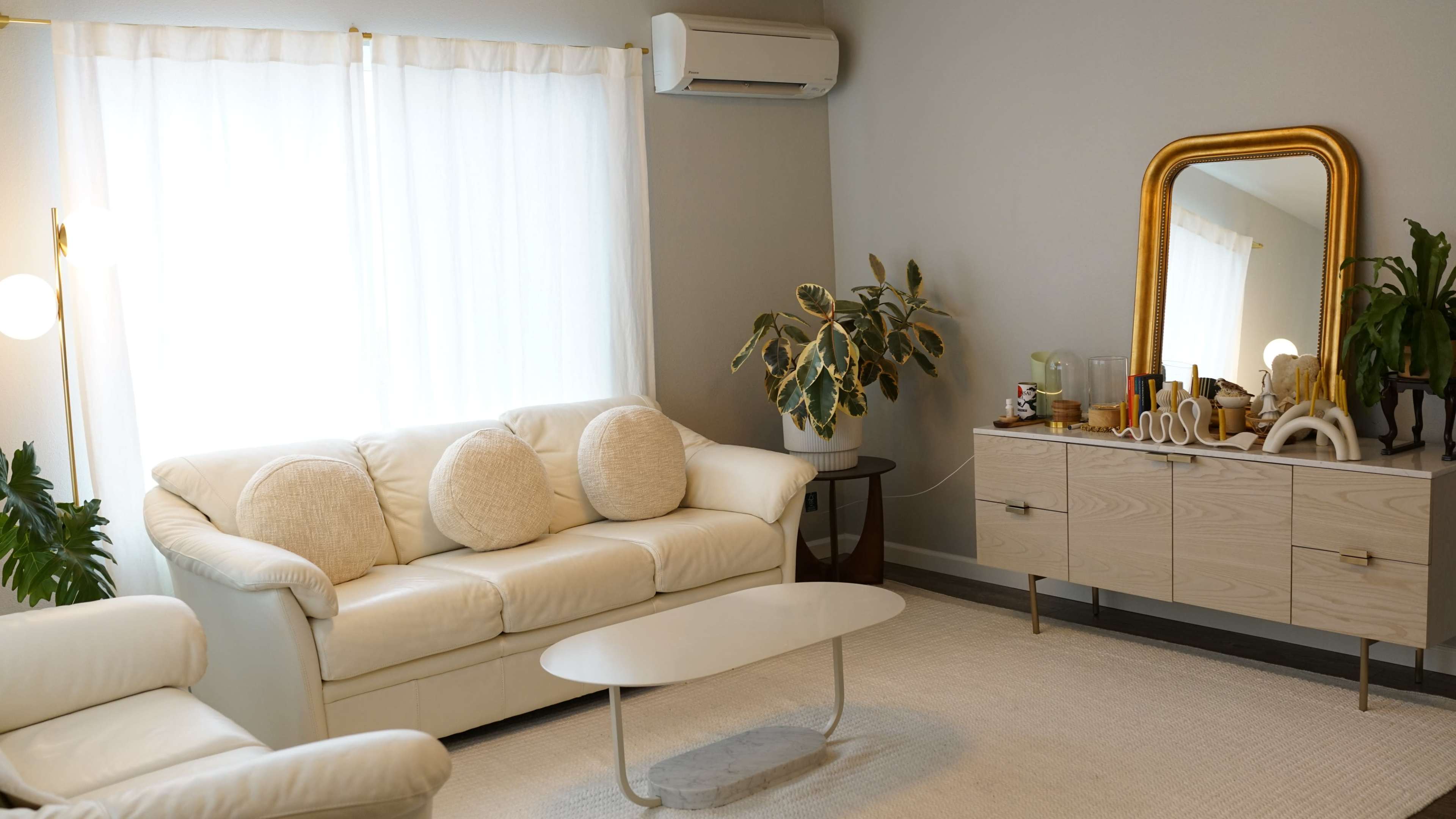 A living room with a light-colored sofa, a white coffee table, and a wooden sideboard, accompanied by a large mirror and a potted plant.