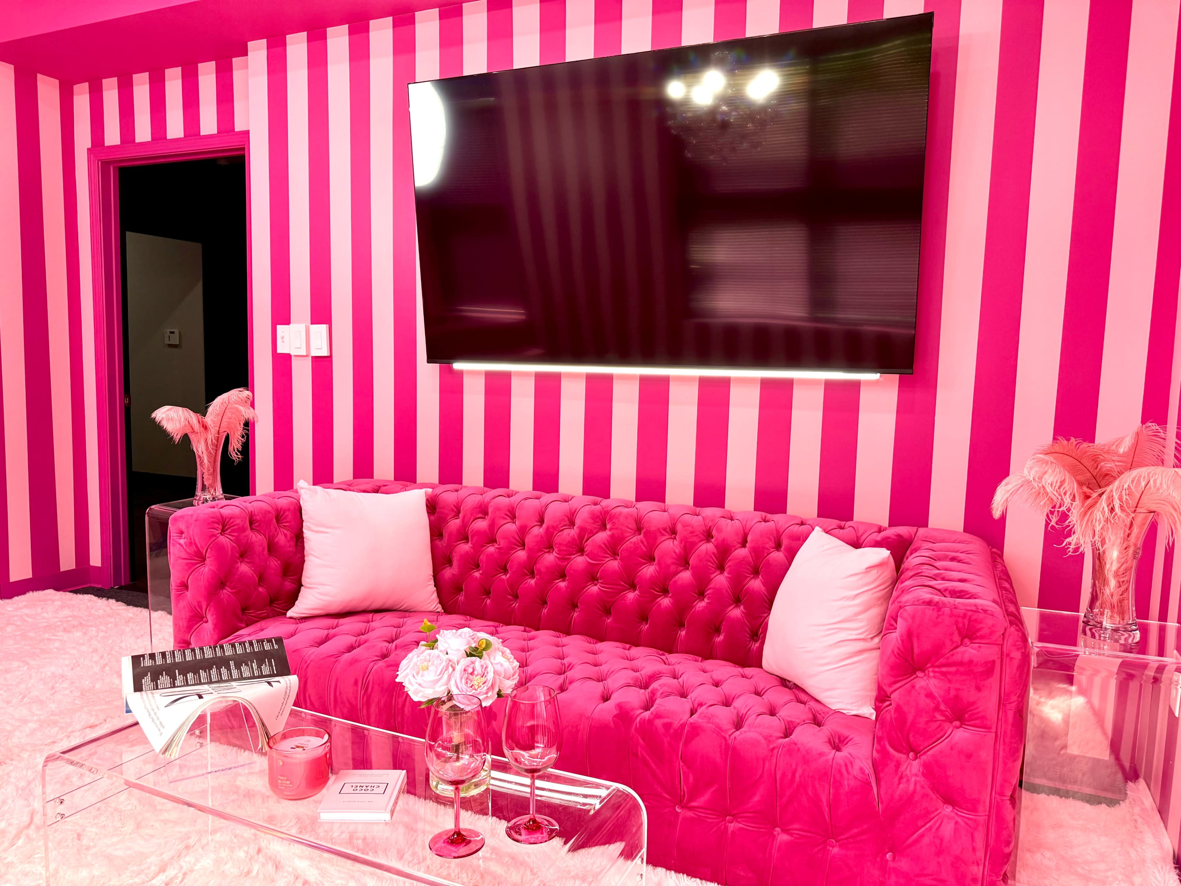 The image shows a living room with a bright pink striped wall, a tufted pink sofa, a wall-mounted television, and decorative items like flowers and an elegant table.