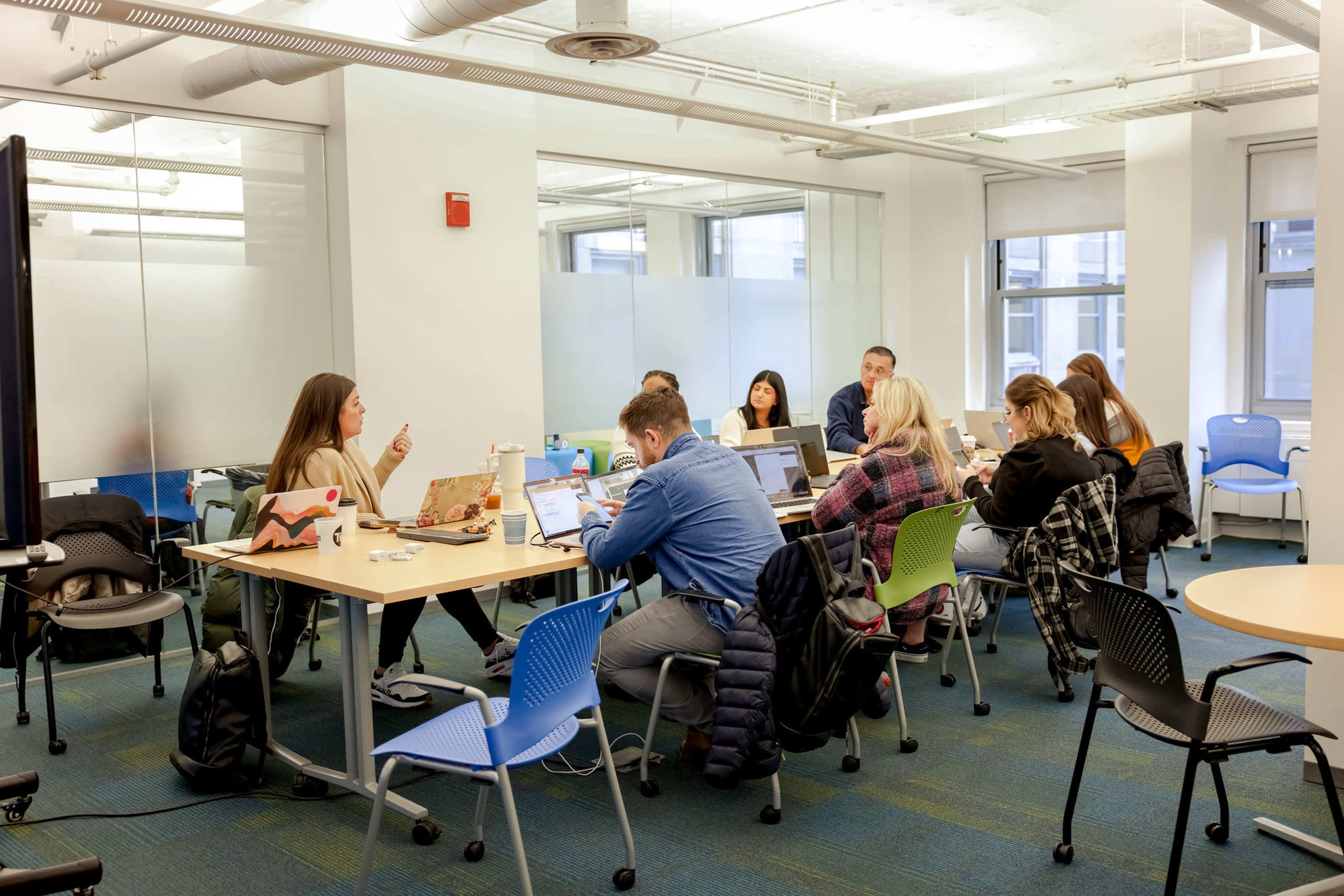 A group of people is engaged in a discussion around a table in a bright, modern meeting room with laptops open in front of them.