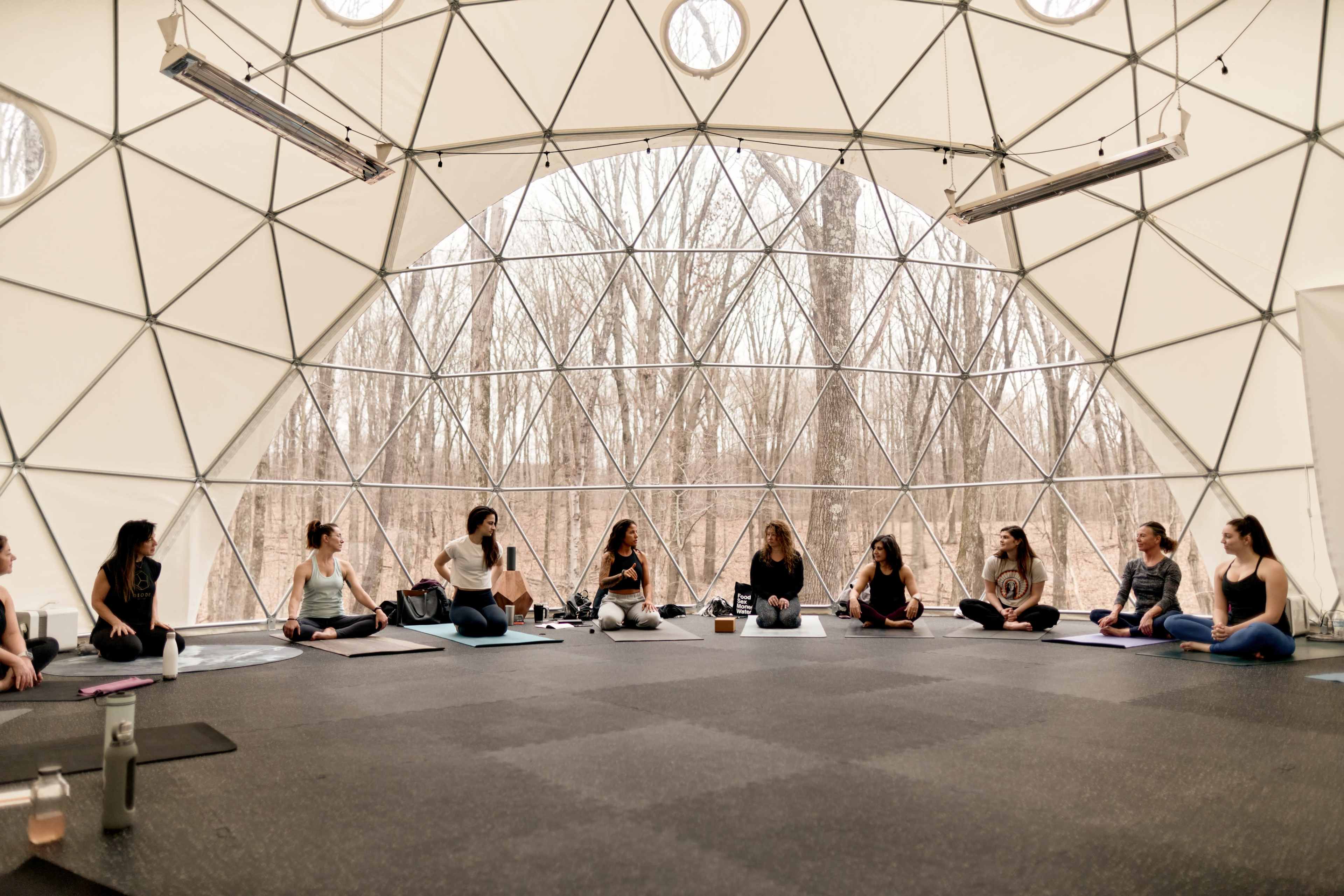 A group of people sits in a circle on mats inside a geodesic dome, surrounded by large windows overlooking a forest.