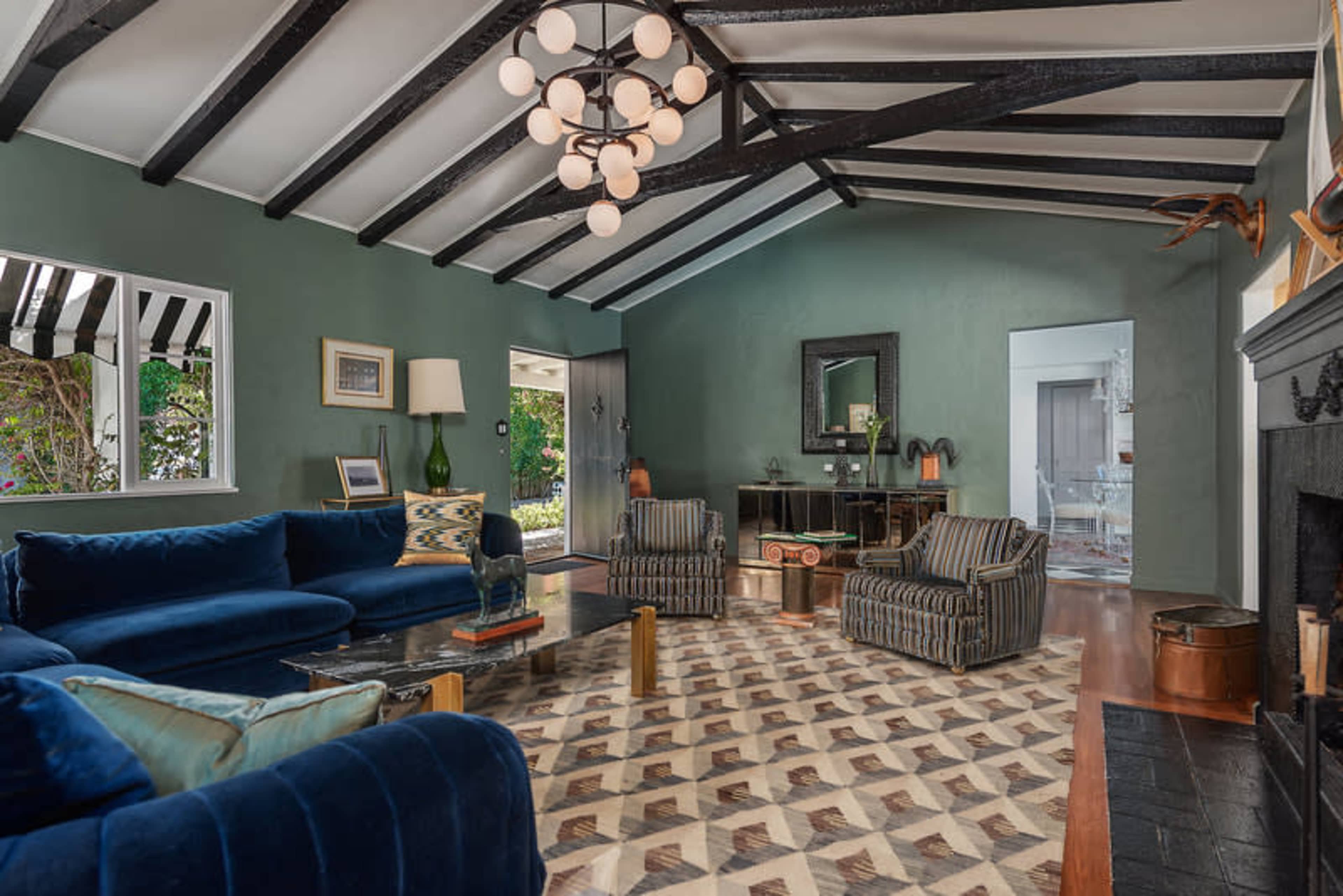 A living room with a vaulted ceiling, featuring a blue sofa and striped armchairs, a patterned rug, and large windows letting in natural light.
