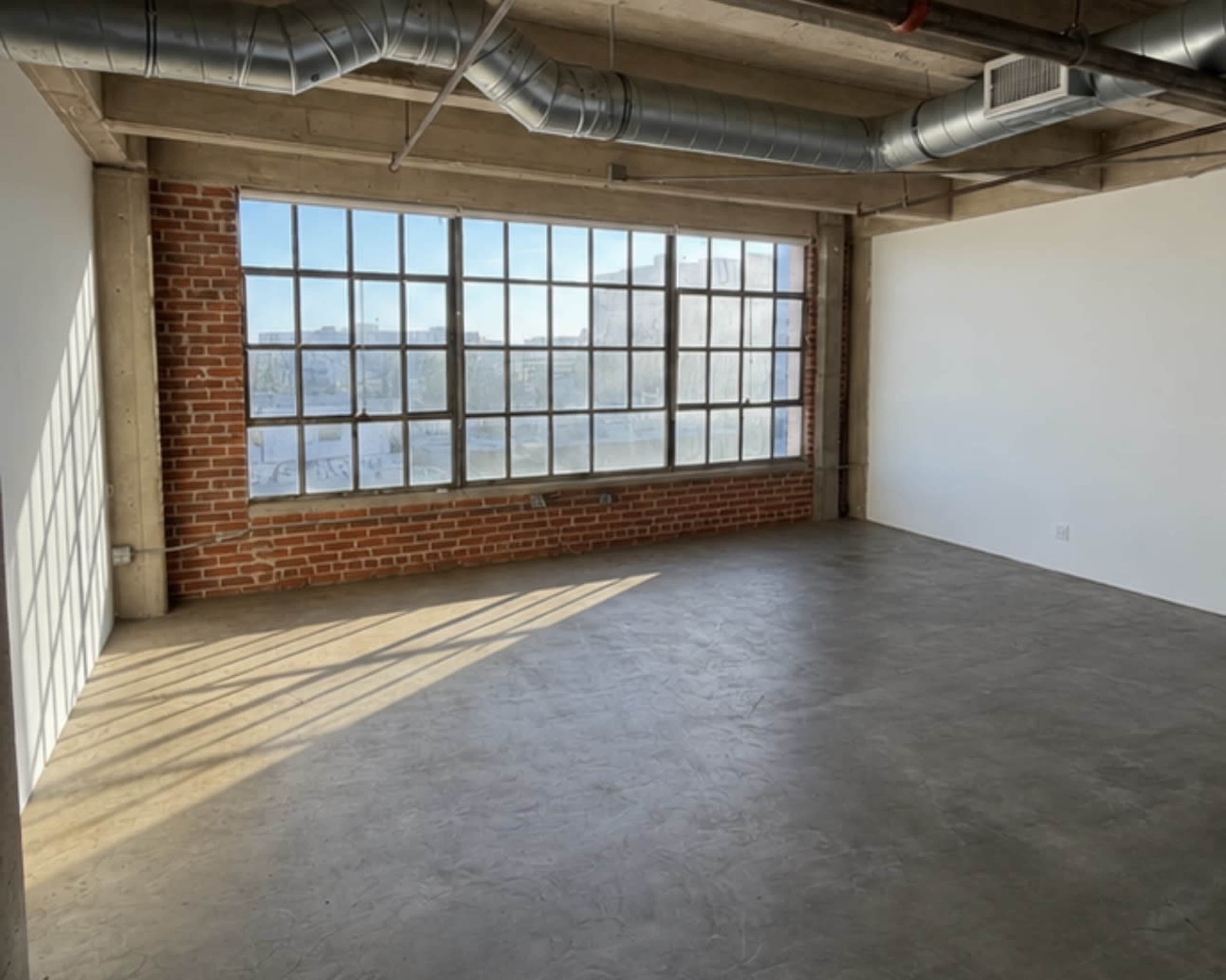 The image shows a spacious, industrial-style room with large windows and exposed brick walls.