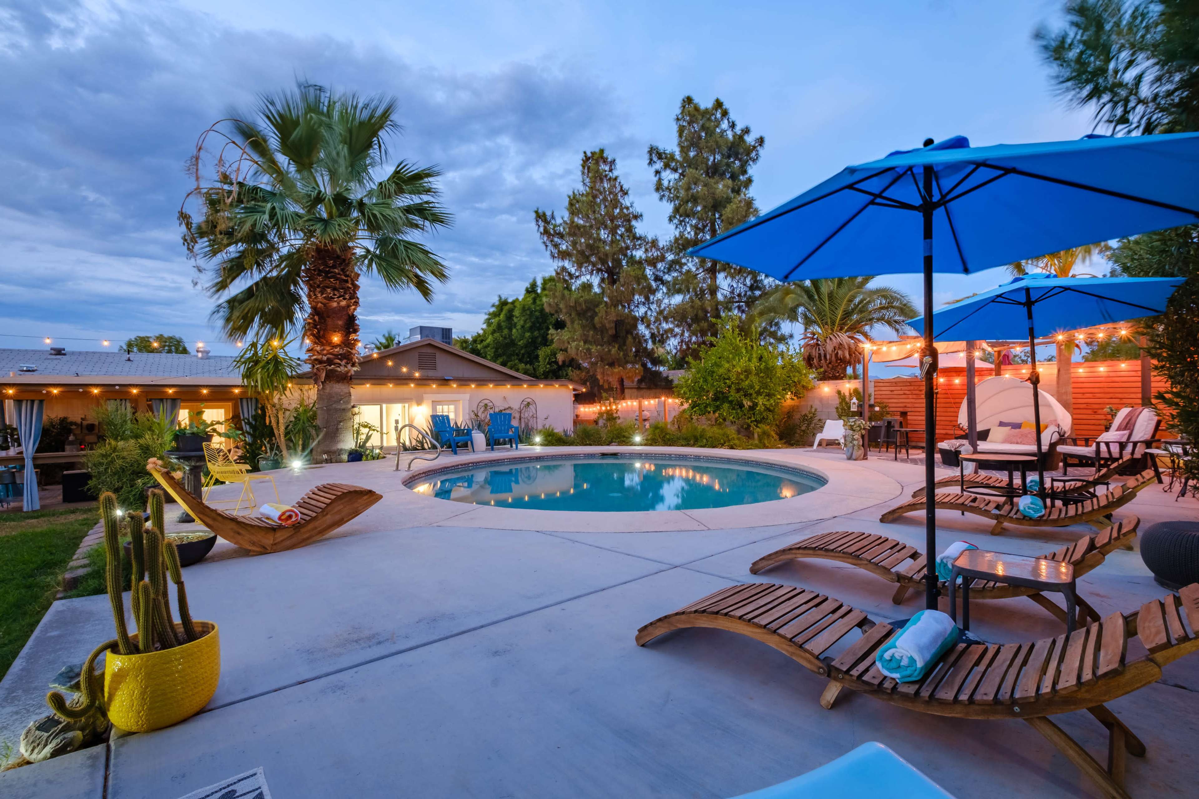 A spacious backyard features a swimming pool surrounded by lounge chairs and large umbrellas, with palm trees and string lights enhancing the outdoor setting.