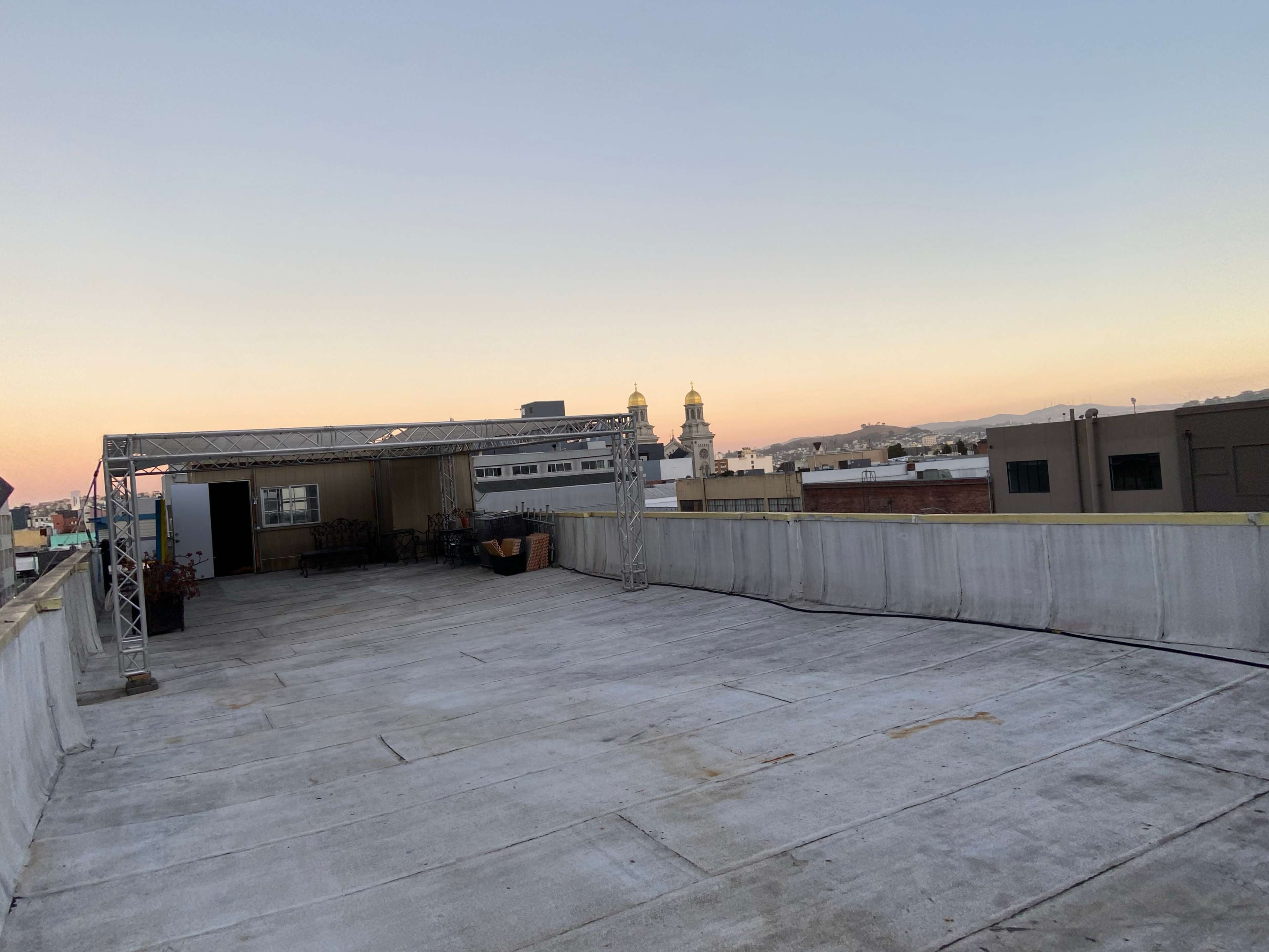 A flat rooftop space with a metal structure and a view of distant buildings and a church steeple at sunset.