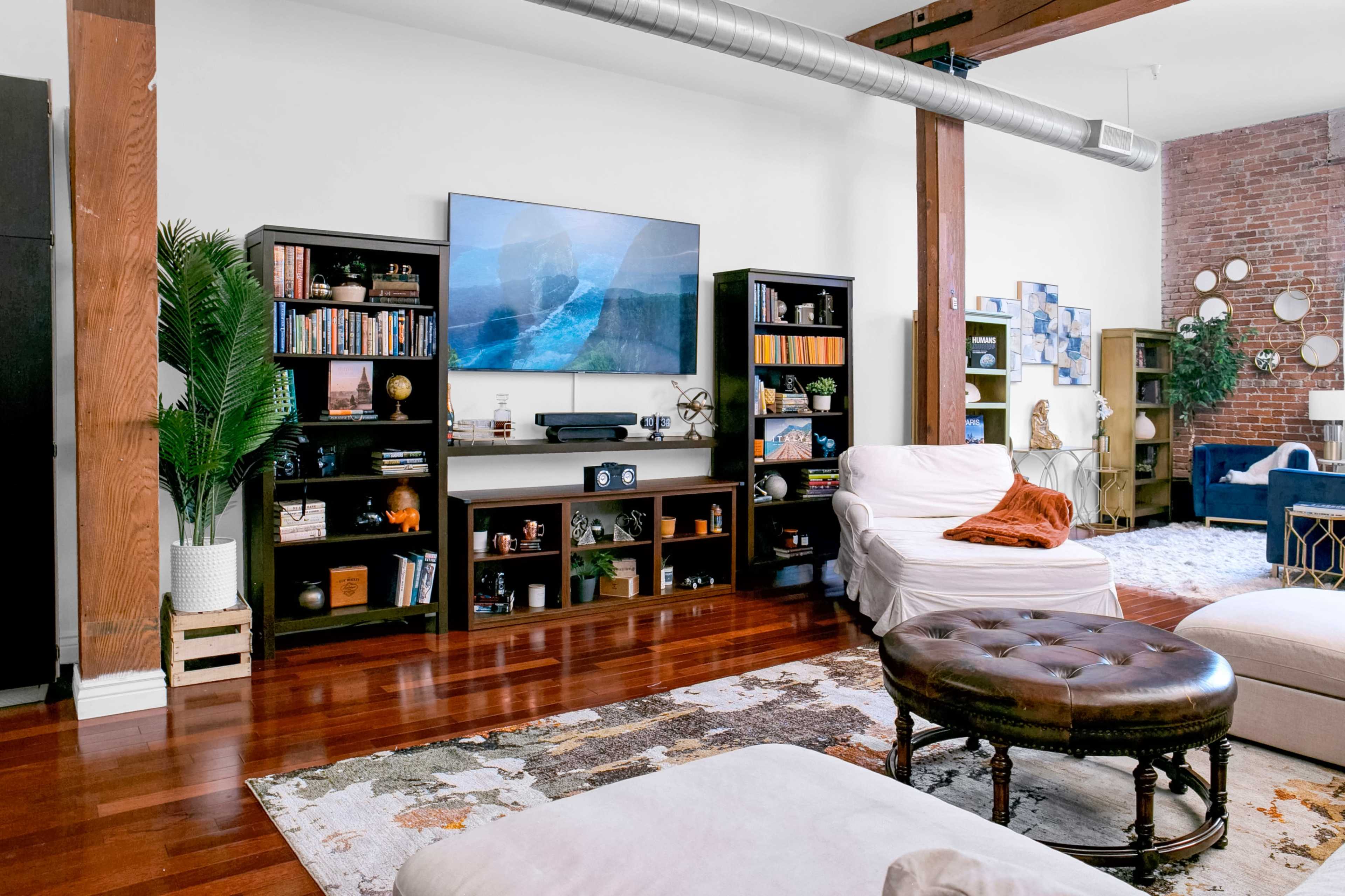 The image shows a modern living room with wooden floors, bookshelves filled with books and decor, and a large television mounted on the wall.