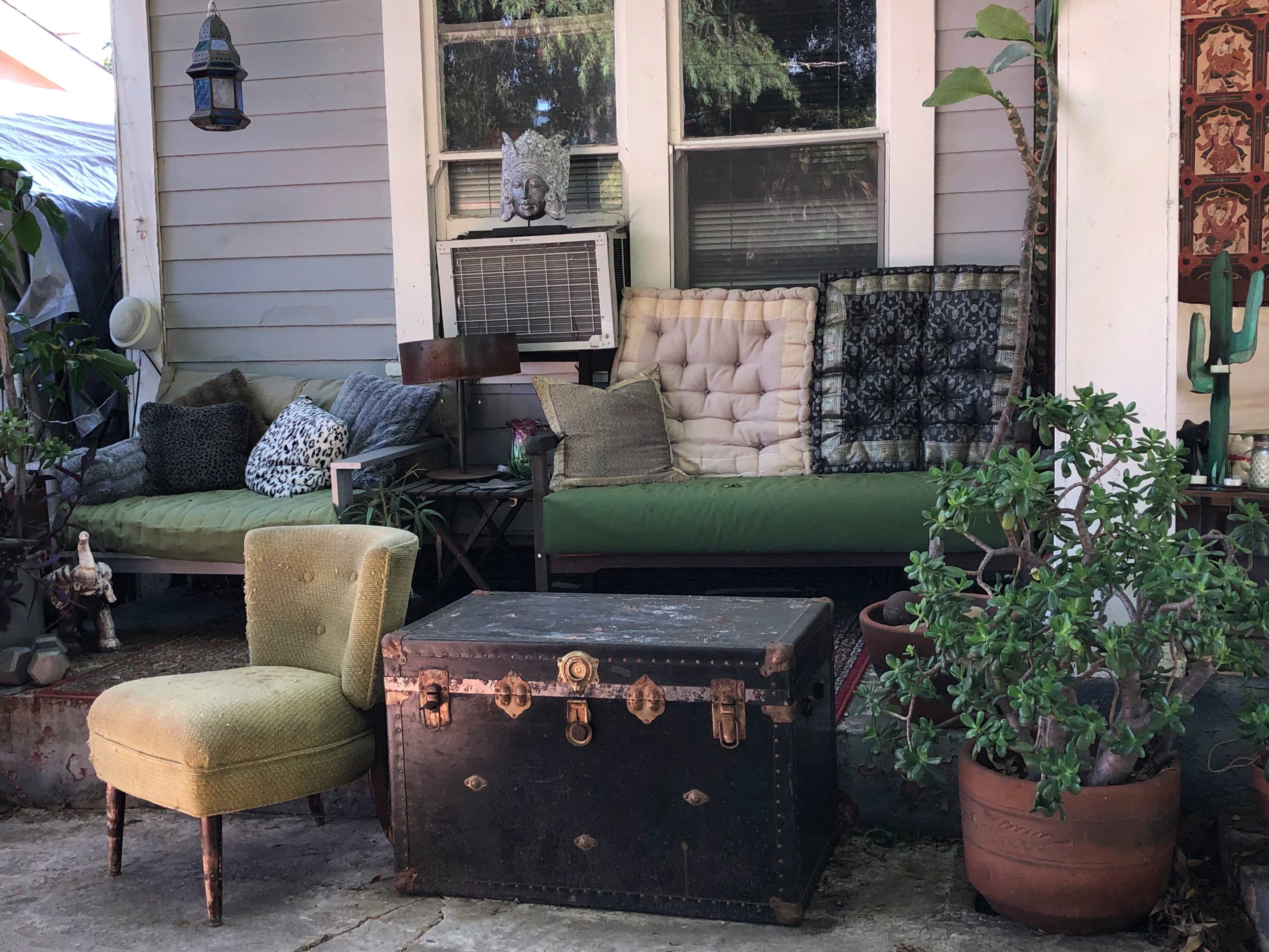 The scene features a porch with a green couch adorned with various cushions, a wooden chair, a black trunk, and potted plants.