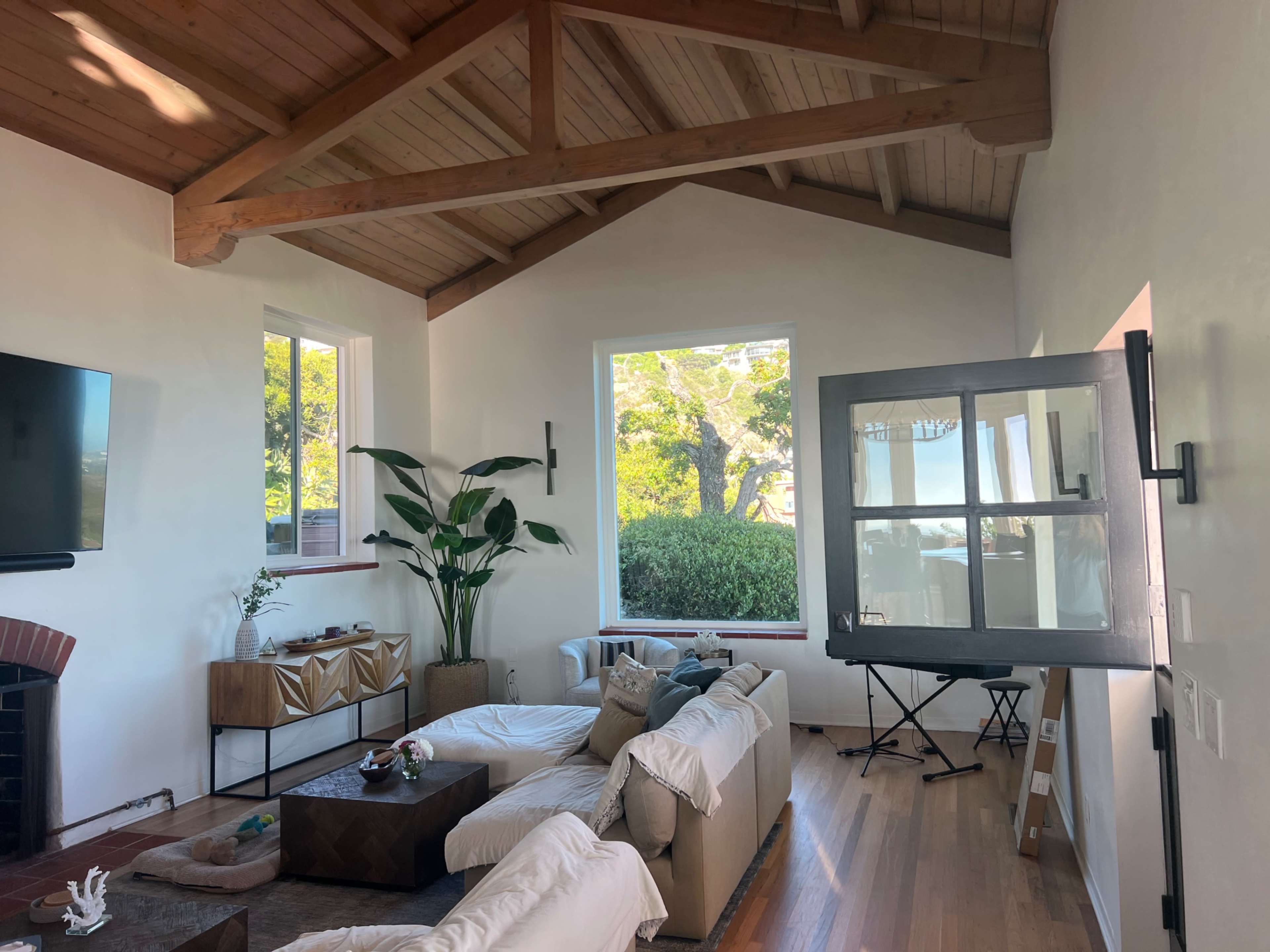 The image shows a spacious living room with high wooden ceilings, large windows, a couch, and a decorative plant.