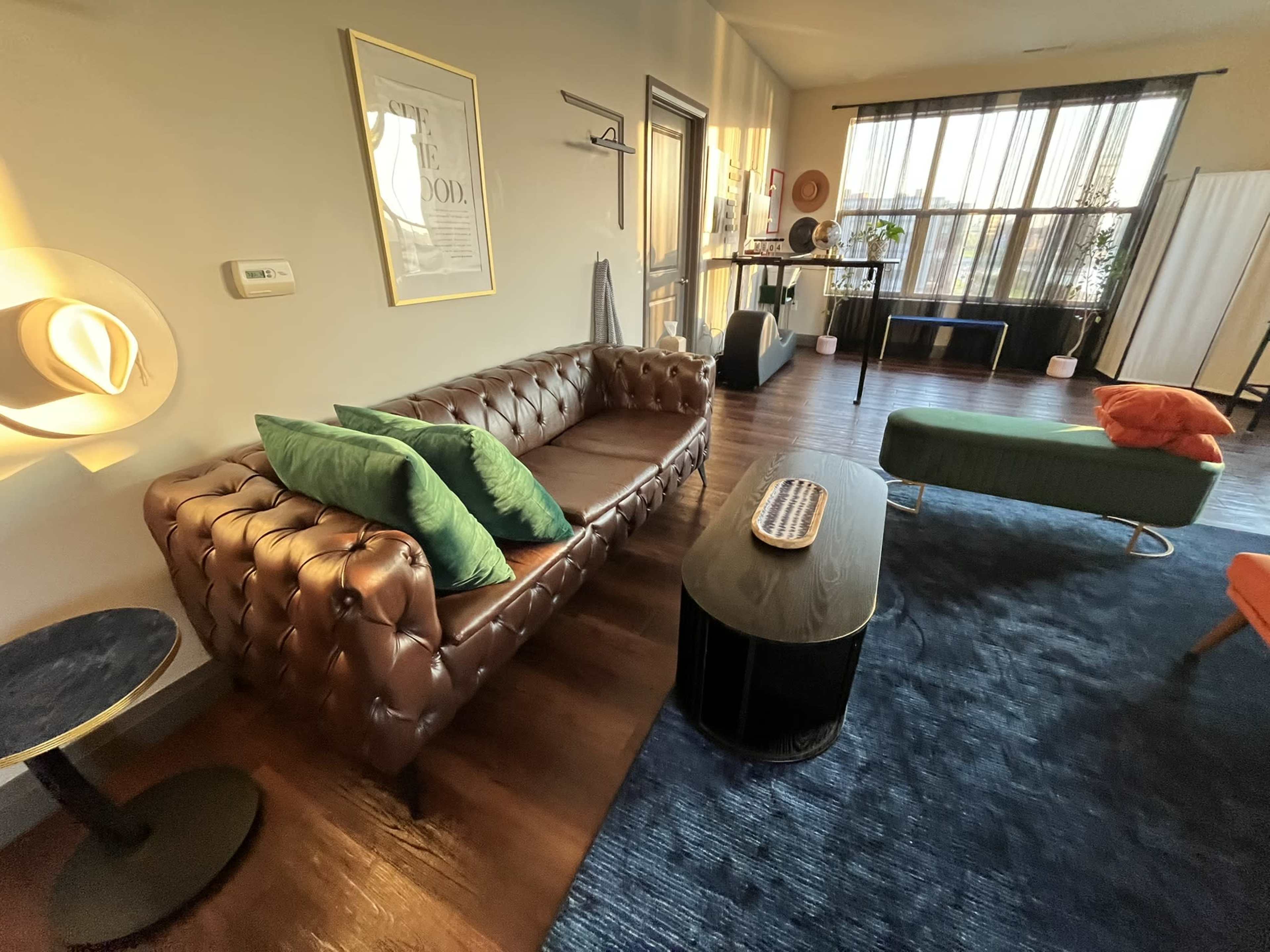 The image depicts a stylish living room featuring a brown leather chesterfield sofa with green cushions, a black coffee table, and various pieces of furniture arranged around a large blue rug.