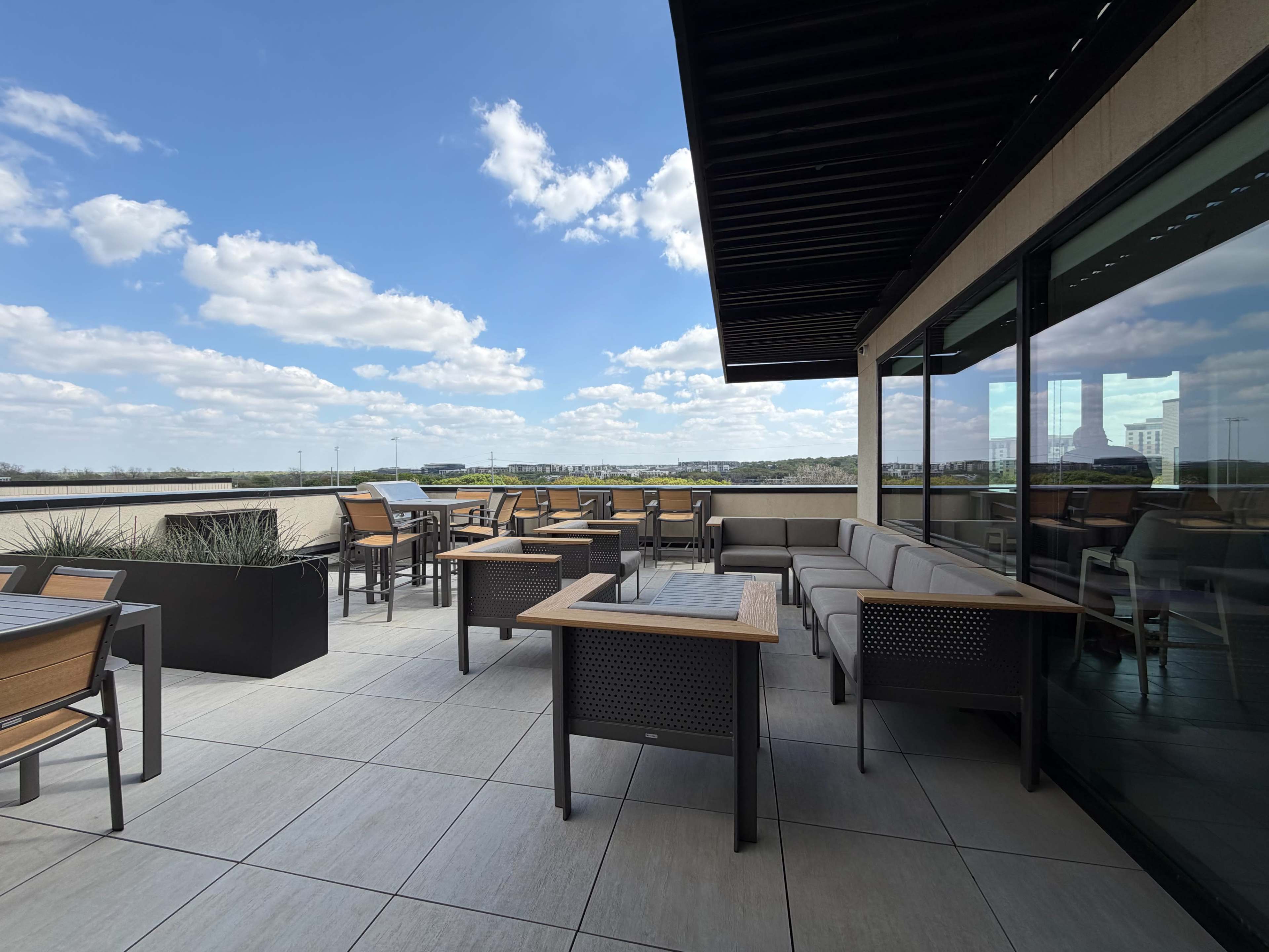 The image shows an outdoor seating area on a rooftop, featuring tables and chairs with a view of the landscape under a partly cloudy sky.