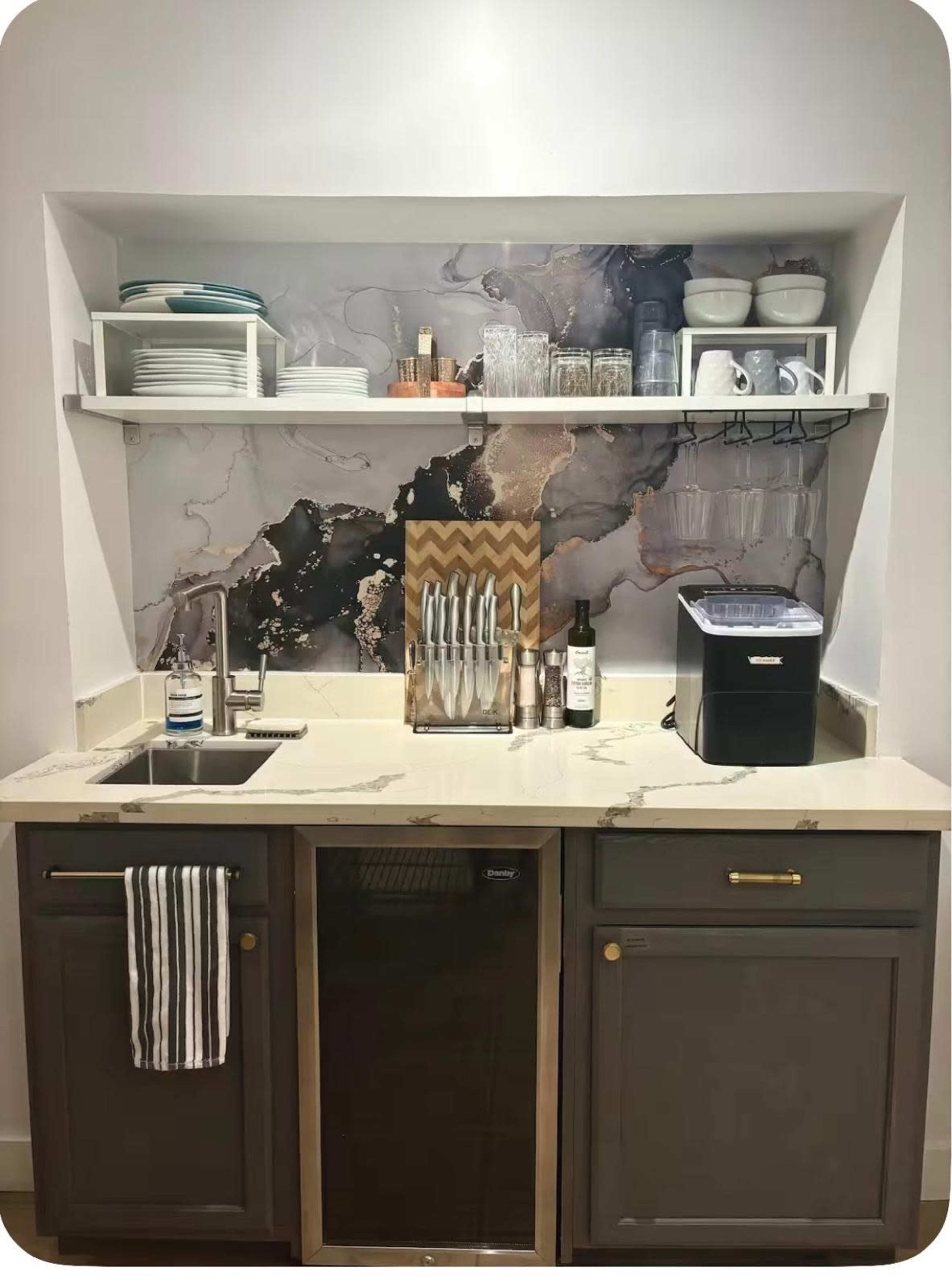 The image shows a small kitchen space with a marble countertop, dark cabinets, a sink, a mini-bar refrigerator, an ice maker, glassware, and plates on open shelves.