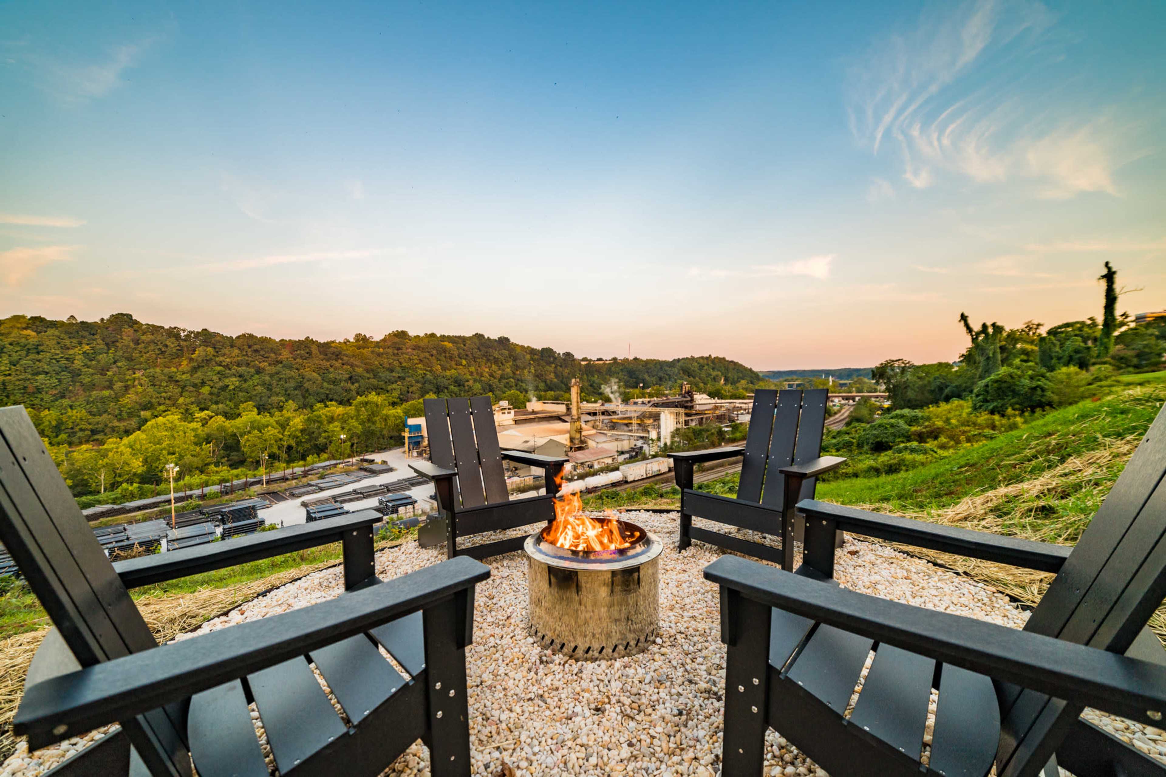 Four black adirondack chairs surround a fire pit on a hillside overlooking a valley with trees and industrial buildings.