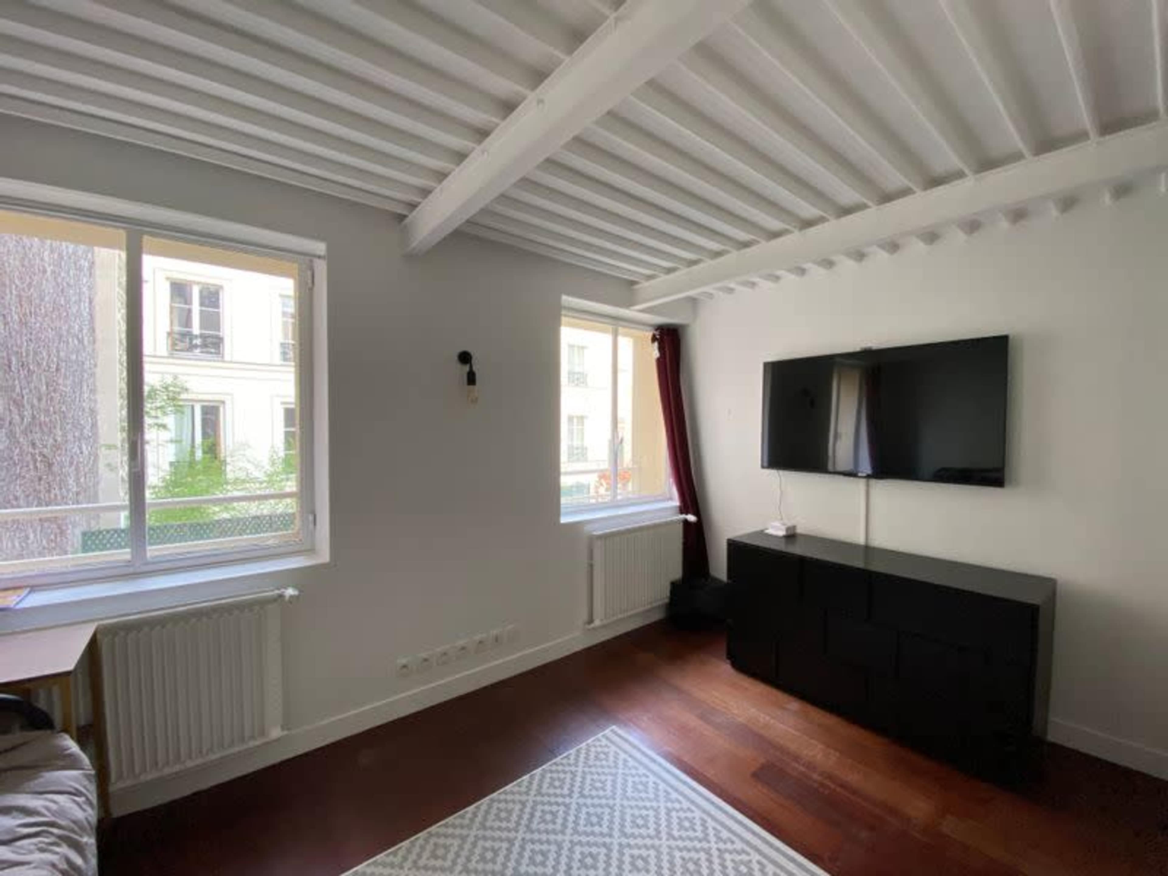 A bright, minimalistic living room features two windows, a wall-mounted television, and a dark media cabinet on hardwood flooring.