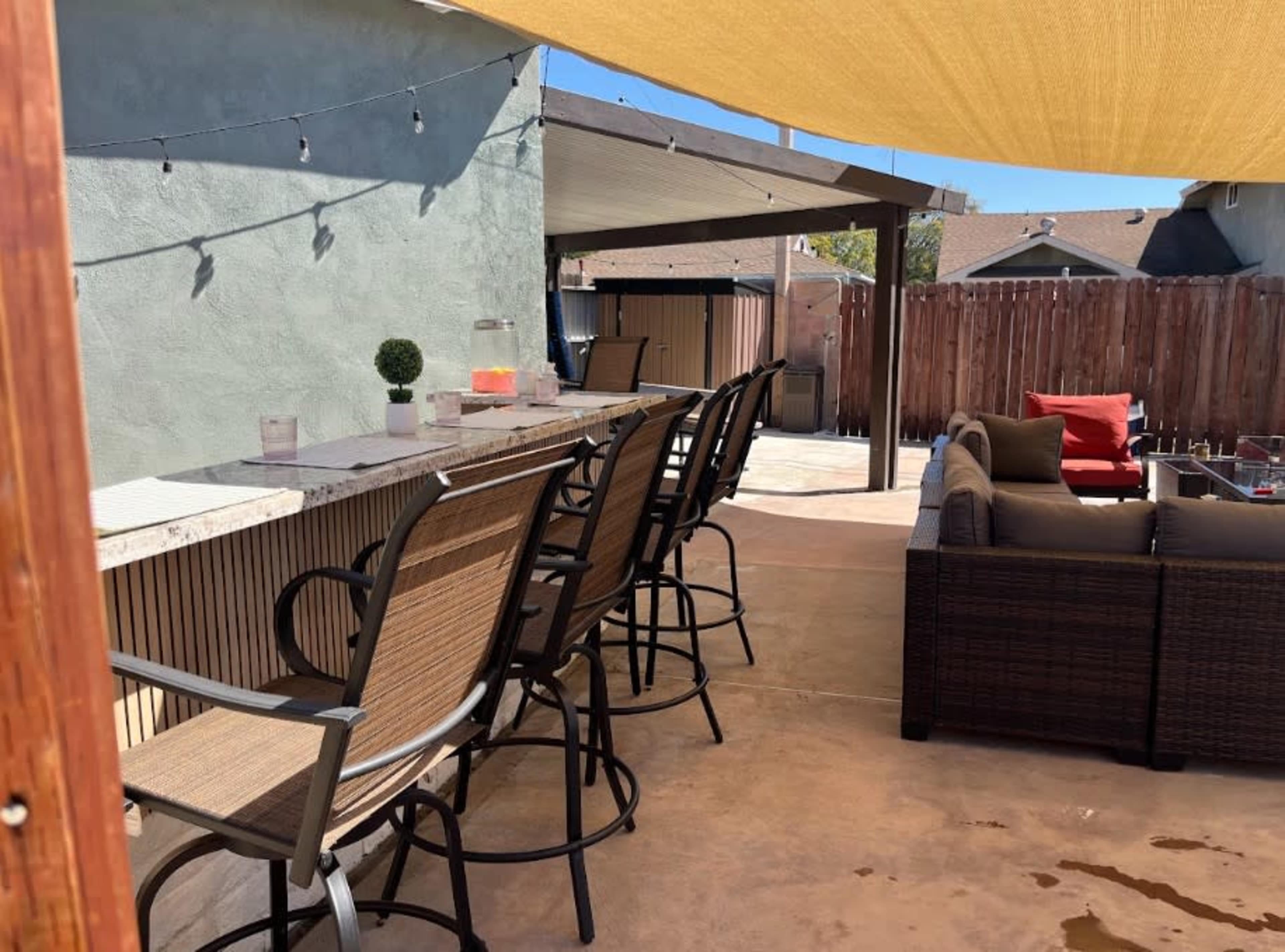 The image shows an outdoor patio area with bar seating, a shaded counter, and a seating arrangement featuring cushions.
