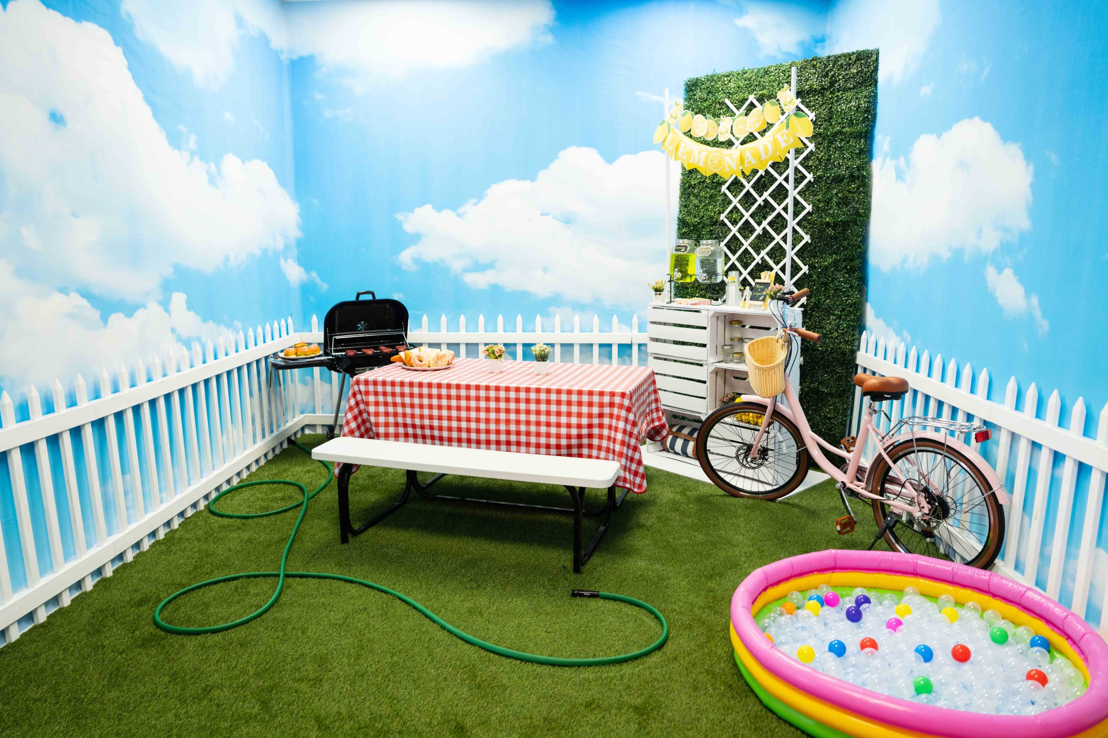 The scene features a picnic setup with a checkered tablecloth, a grill, a bicycle, and a small inflatable pool filled with colorful balls, all enclosed by a white picket fence and decorated with artificial grass and a blue sky backdrop.