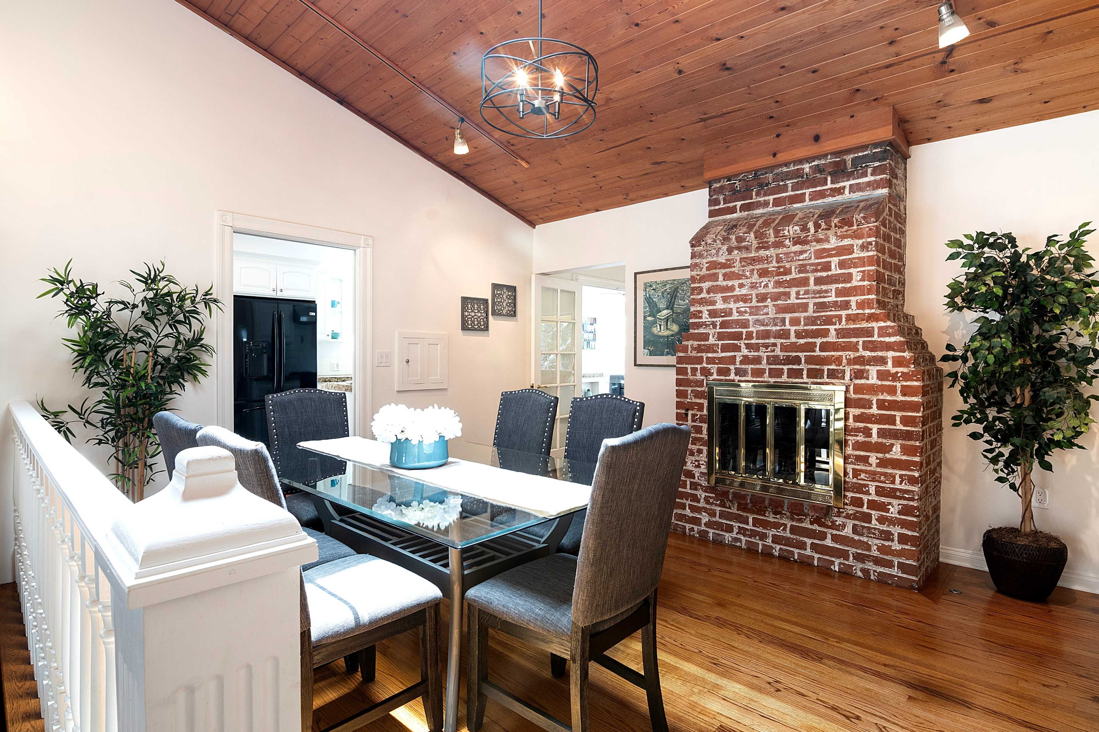 The image shows a dining area with a glass table surrounded by six chairs, a brick fireplace on one wall, and a wooden ceiling.