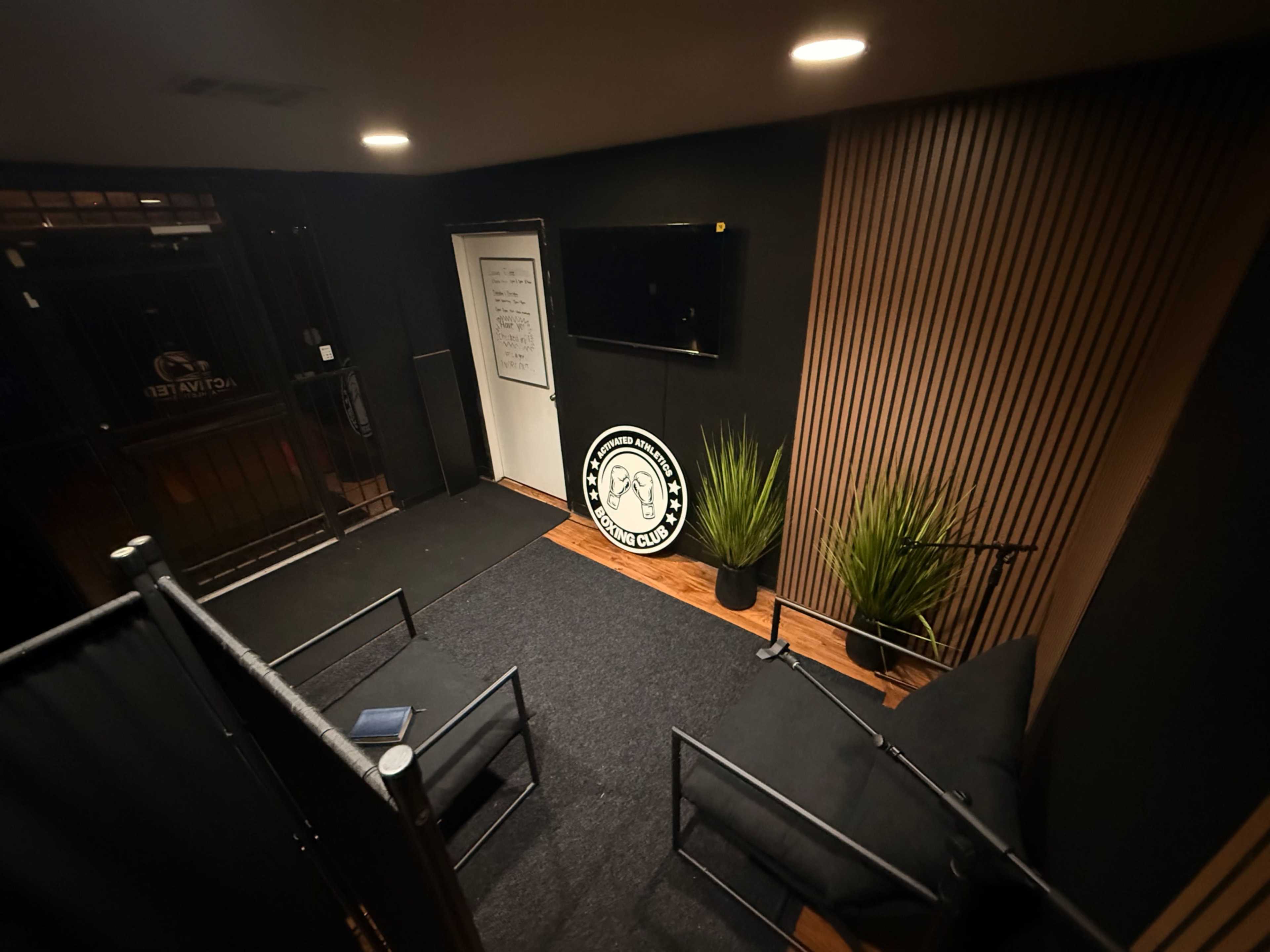 The image shows a small, dark room with a television on the wall, a noticeable round logo on the floor, and minimal furnishings including two black chairs and decorative plants.