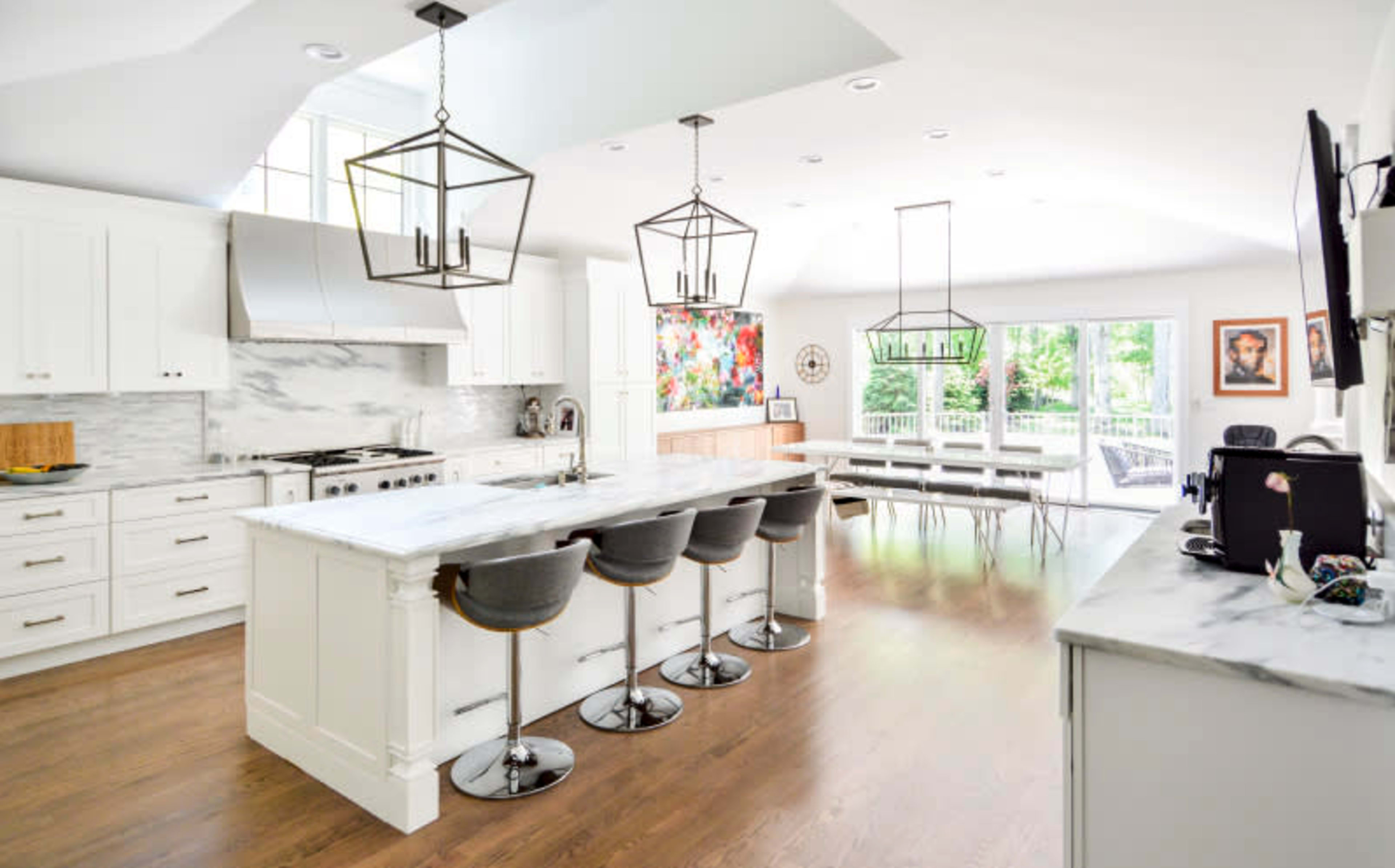 The image shows a bright modern kitchen with a large island, four barstools, and a dining area featuring a long table and large windows overlooking a patio.
