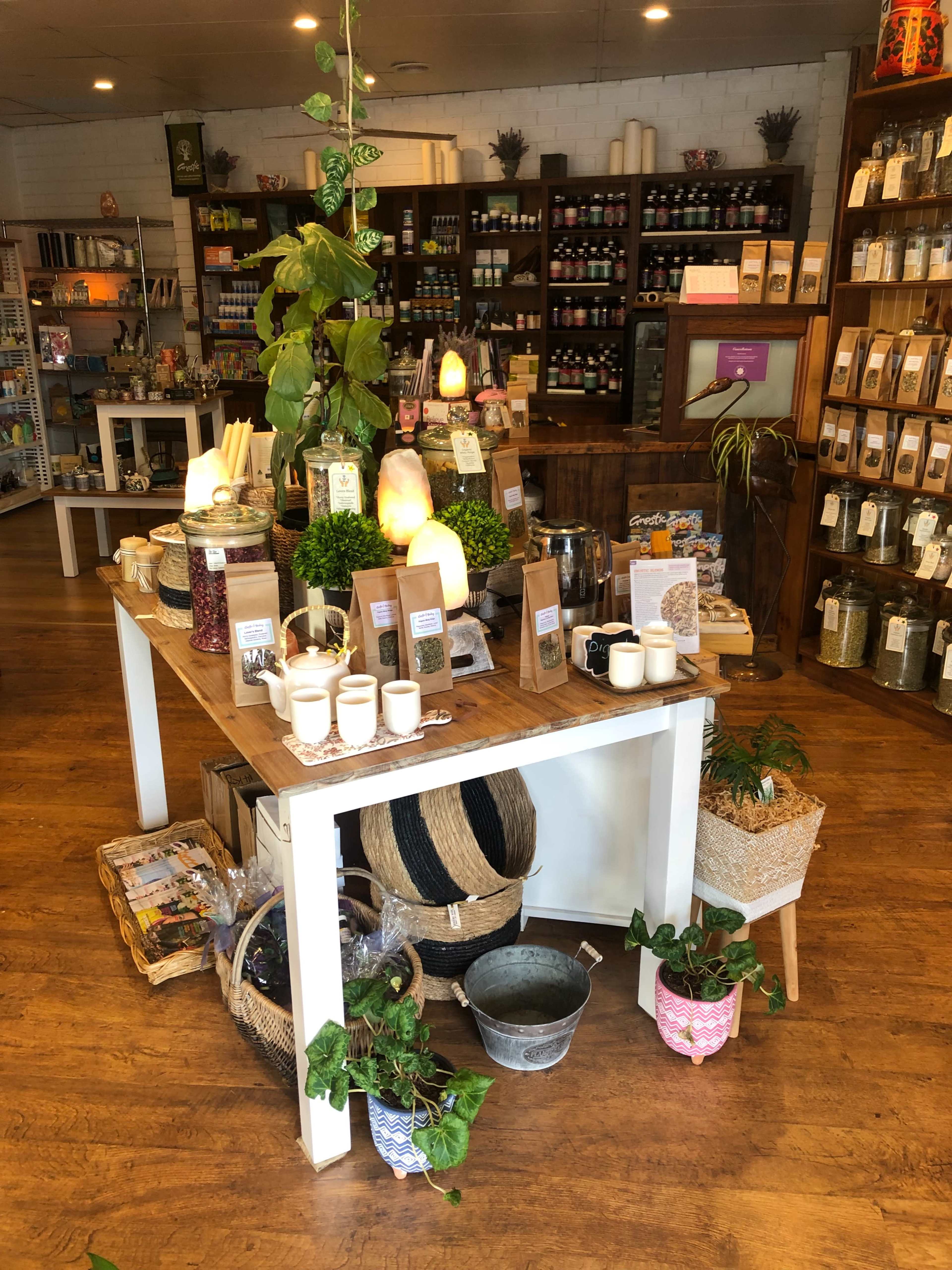 The image shows a well-organized display table in a shop with various products, including jars, candles, and plants, surrounded by shelves filled with items.
