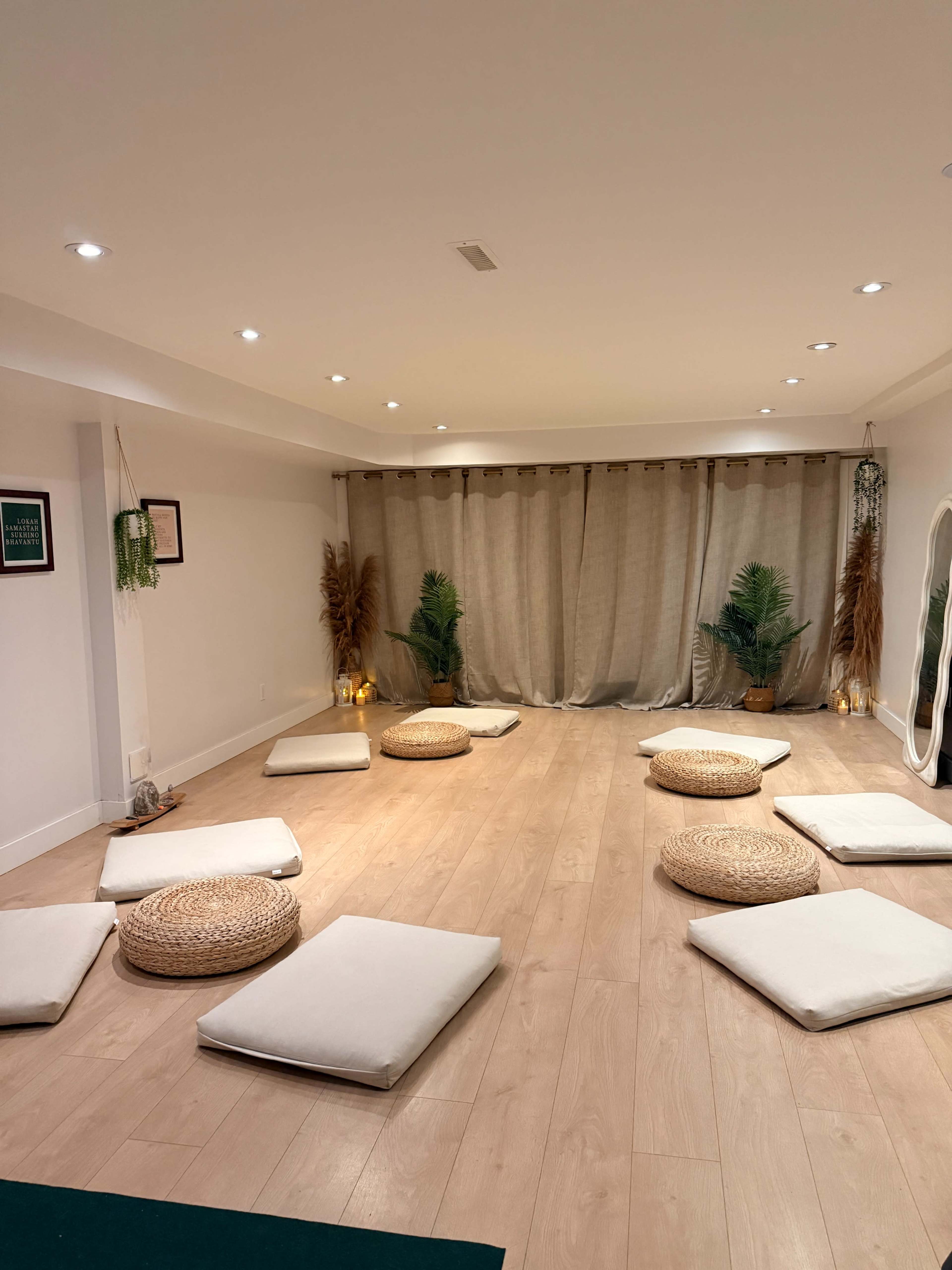 The room features a minimalist design with several floor cushions arranged on a wooden floor, flanked by plants and lit by soft lighting.