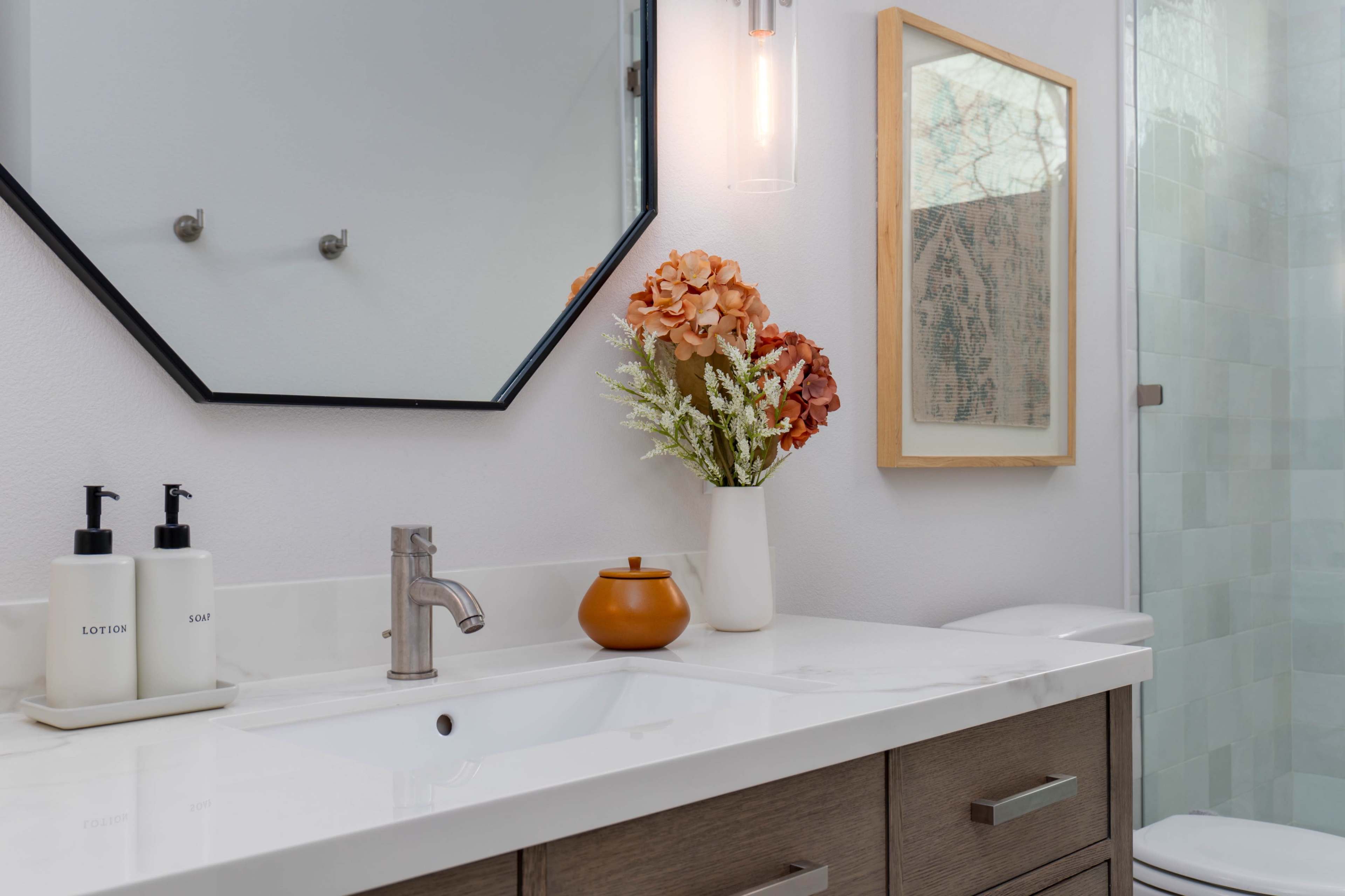 The image shows a modern bathroom featuring a marble countertop with a sink, a vase of flowers, and a framed artwork on the wall.