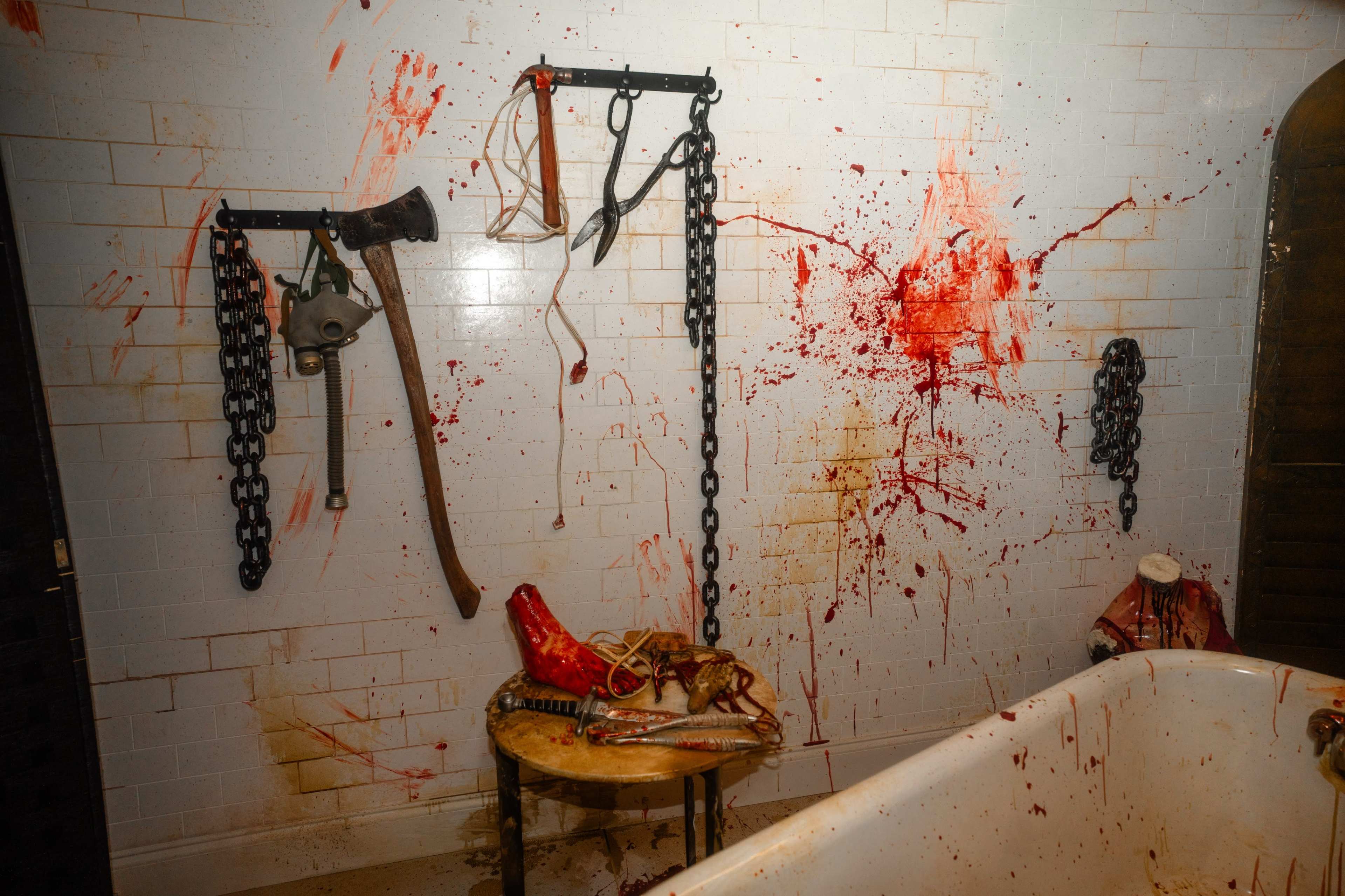 The image depicts a gruesome scene of a bathroom with blood splattered on the walls, an axe, chains, and severed body parts arranged on a table.