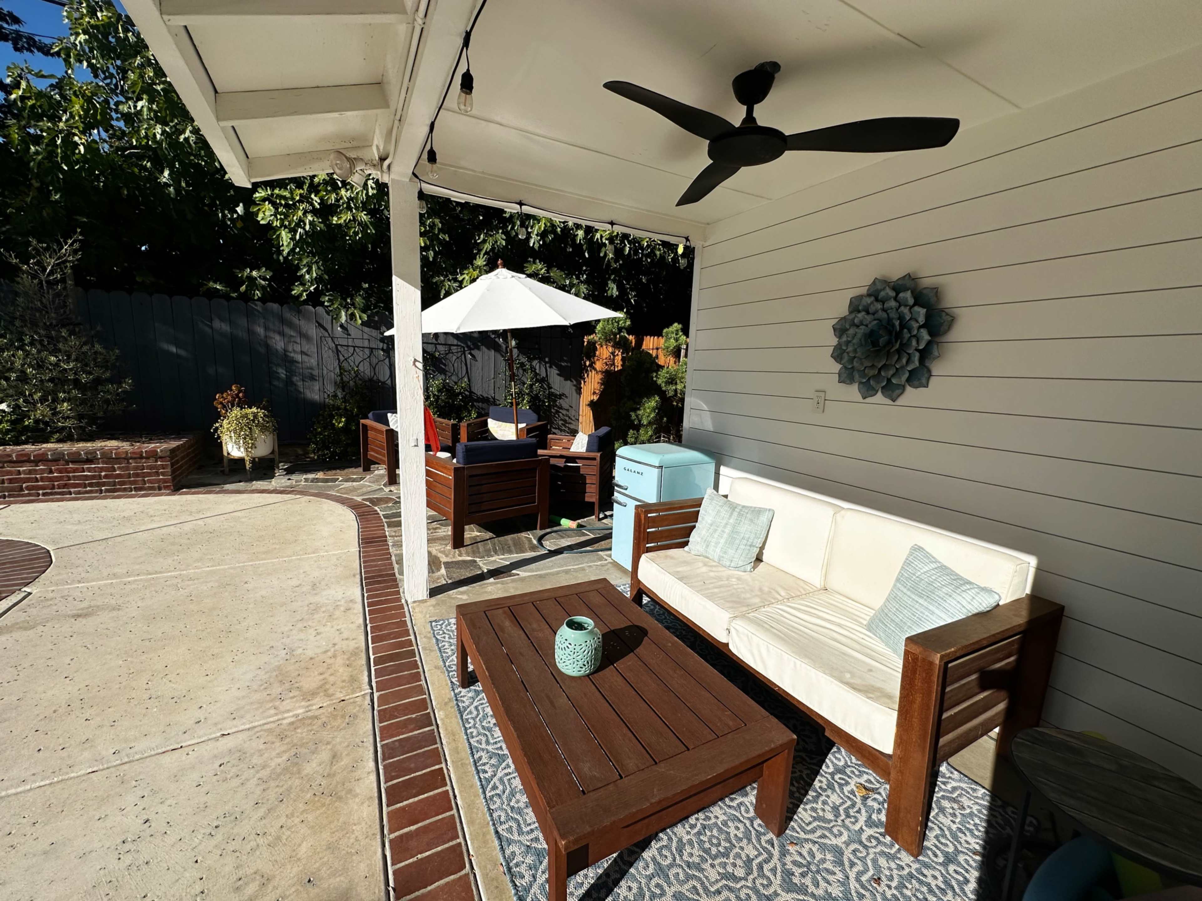 The image shows a cozy outdoor seating area with a white couch, a wooden coffee table, and an umbrella, surrounded by greenery and a brick pathway.
