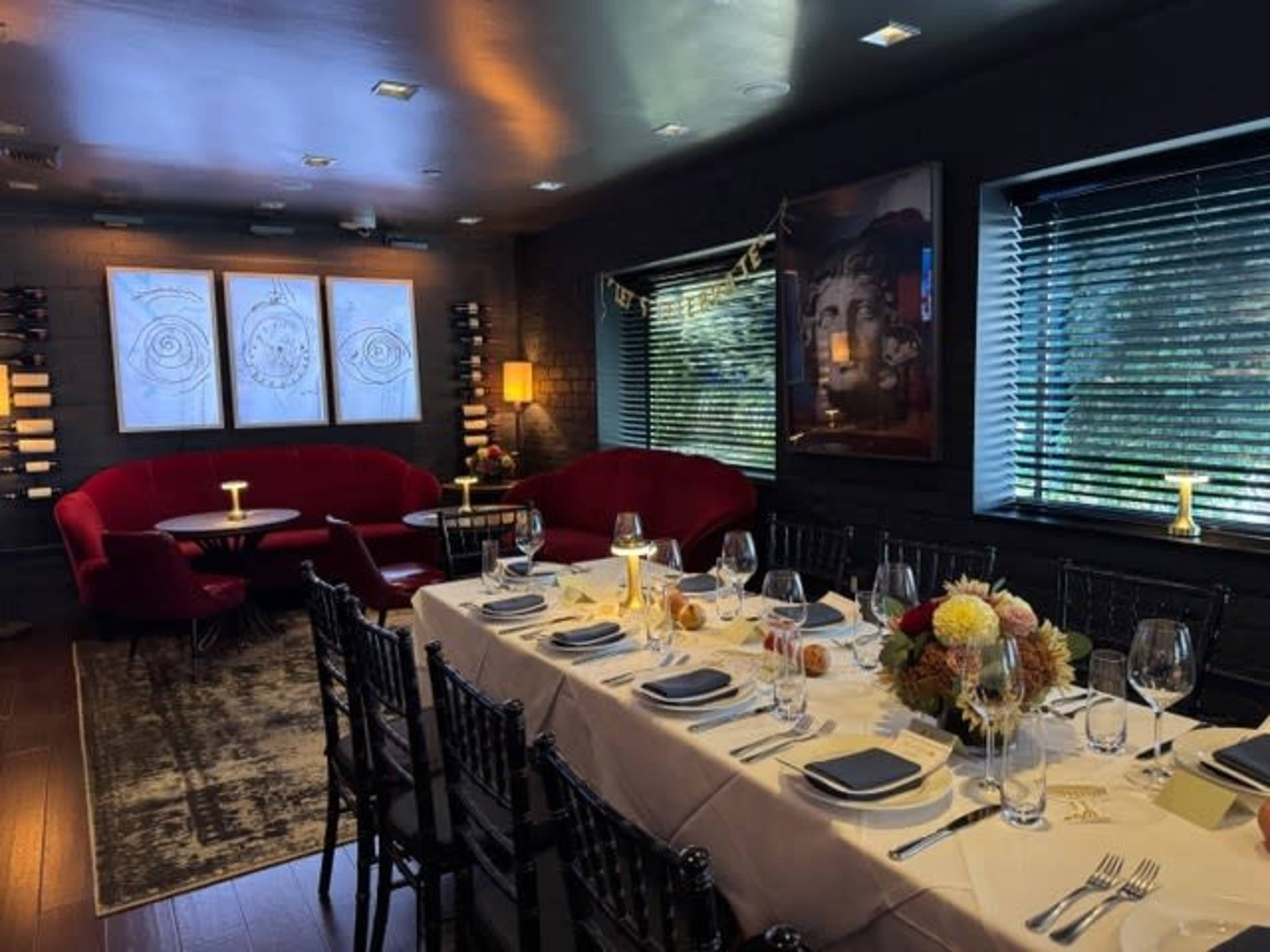 A dining room features a long table set with glassware and plates, flanked by red upholstered seating and illuminated by soft lighting.