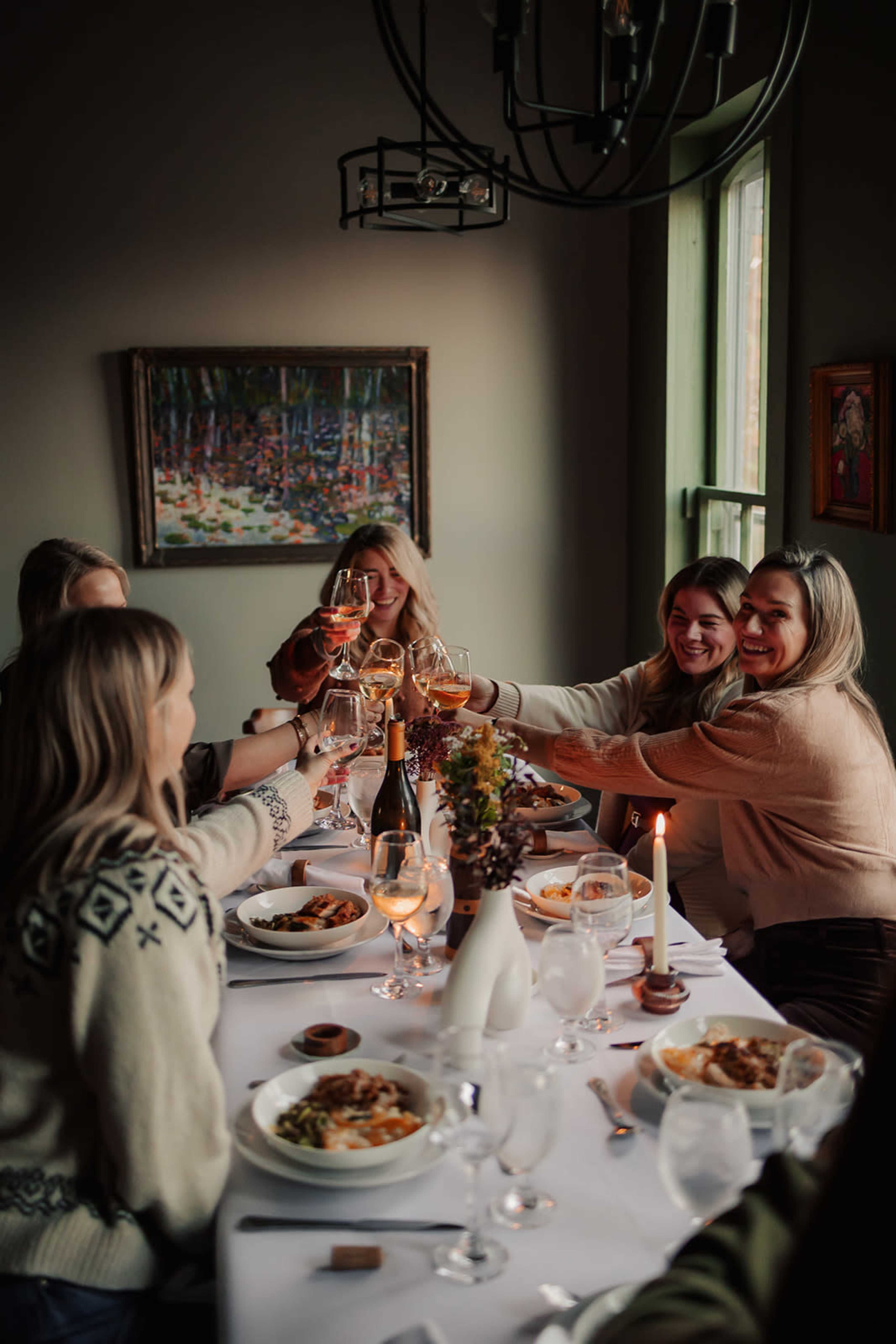 A group of people raises their glasses in a toast at a dining table set with plates of food and candles in a warmly lit room.