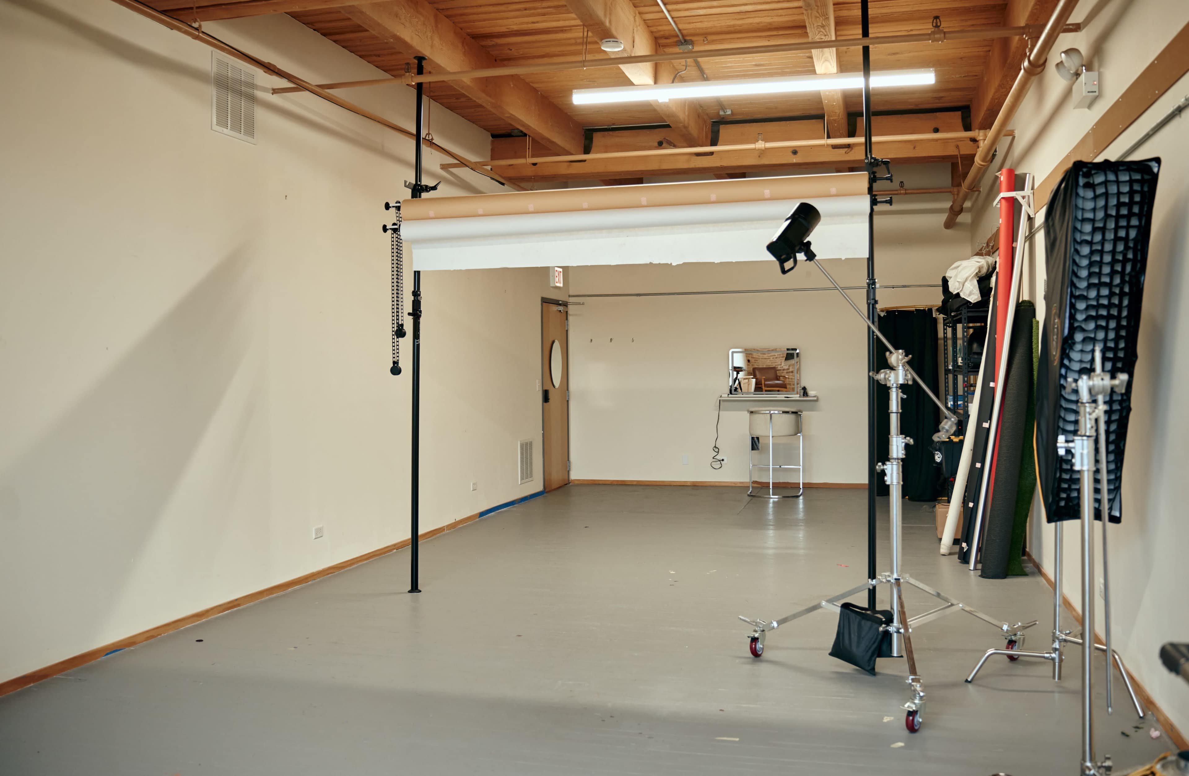 An empty photography studio features a backdrop support system, a lighting setup, and various equipment along the walls.