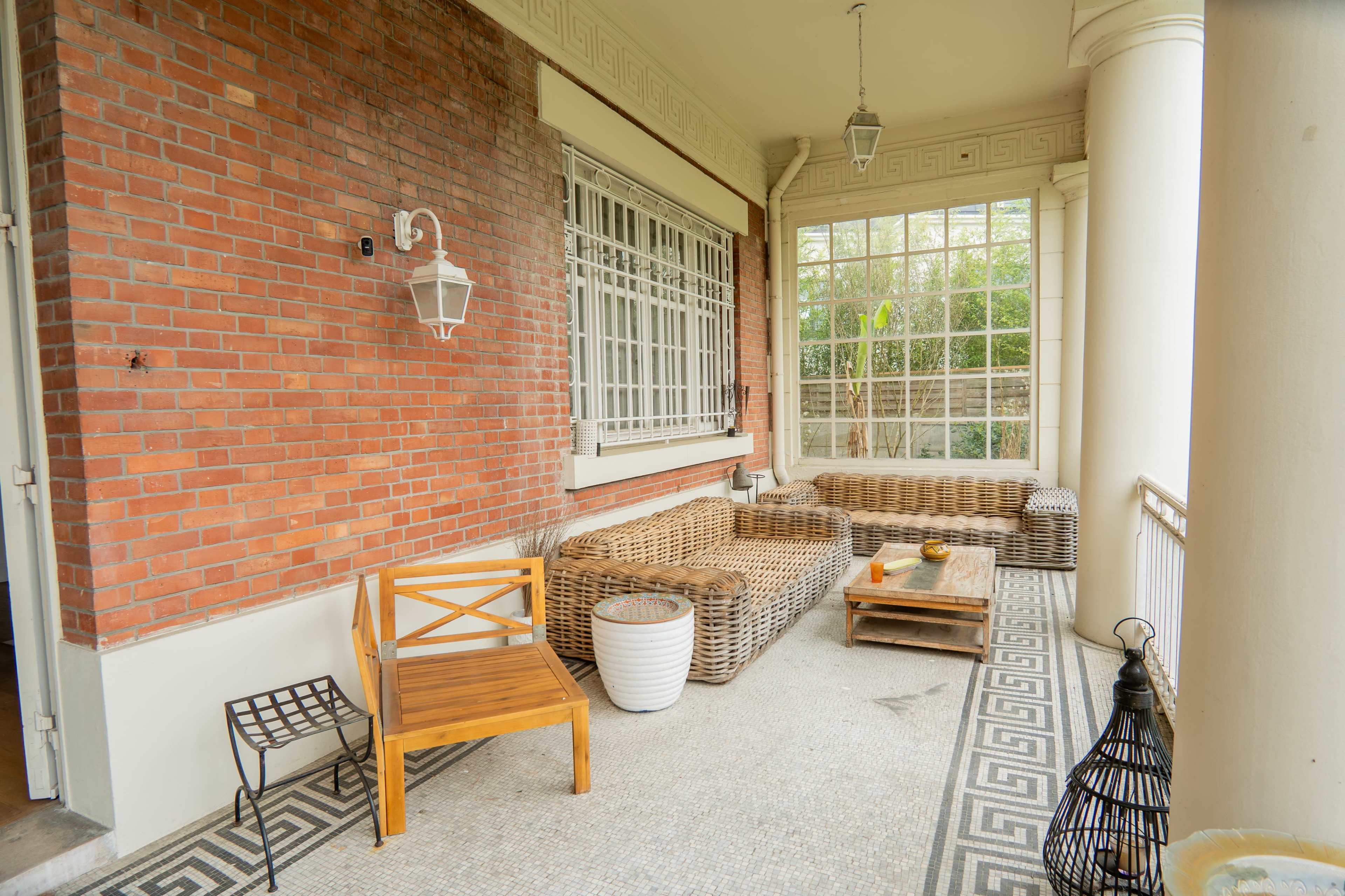 The image shows a spacious porch area with brick walls, wicker seating, and a patterned tiled floor.