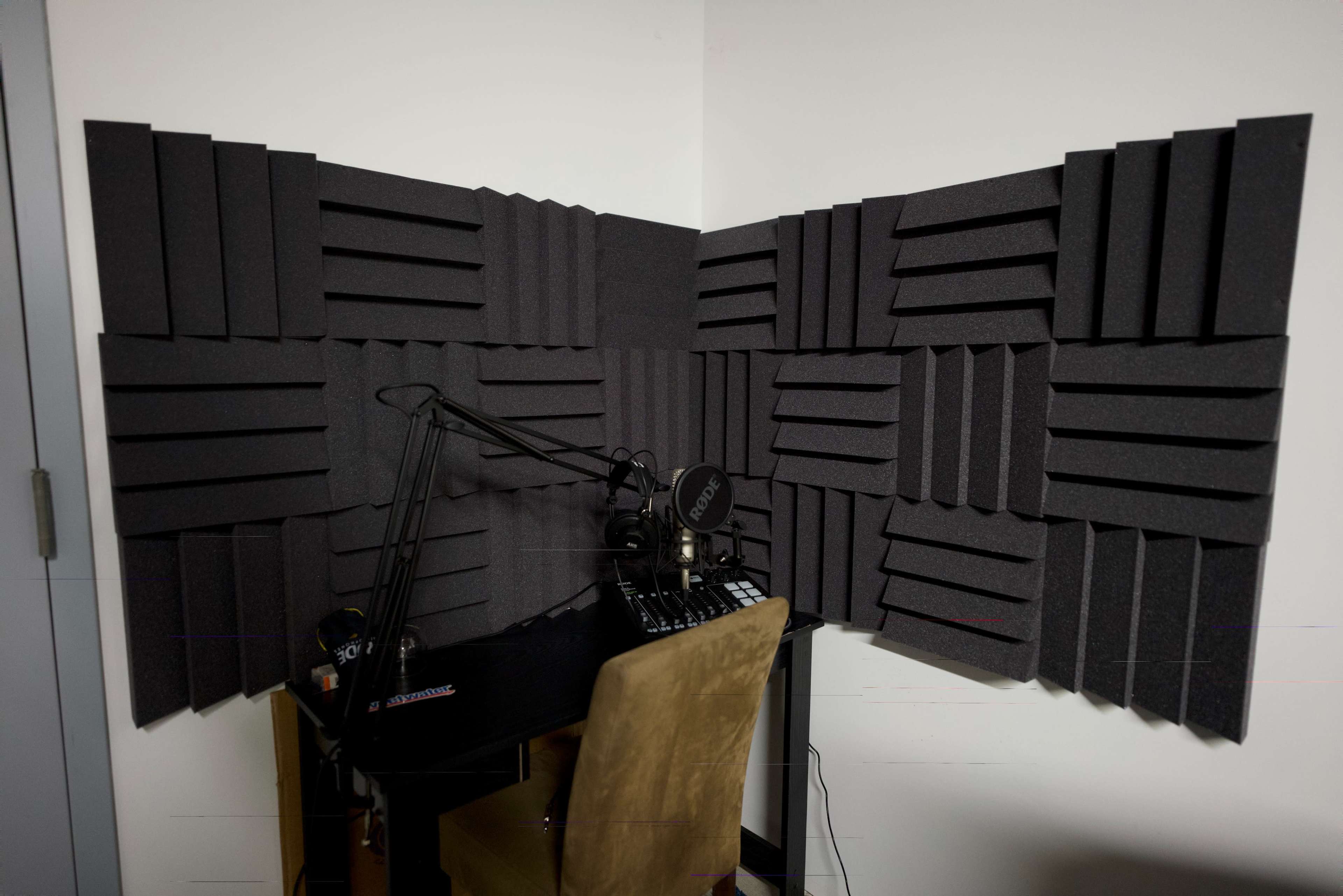 A corner of a room with soundproofing foam panels on the walls, a microphone mounted on a boom arm, and a small desk setup.