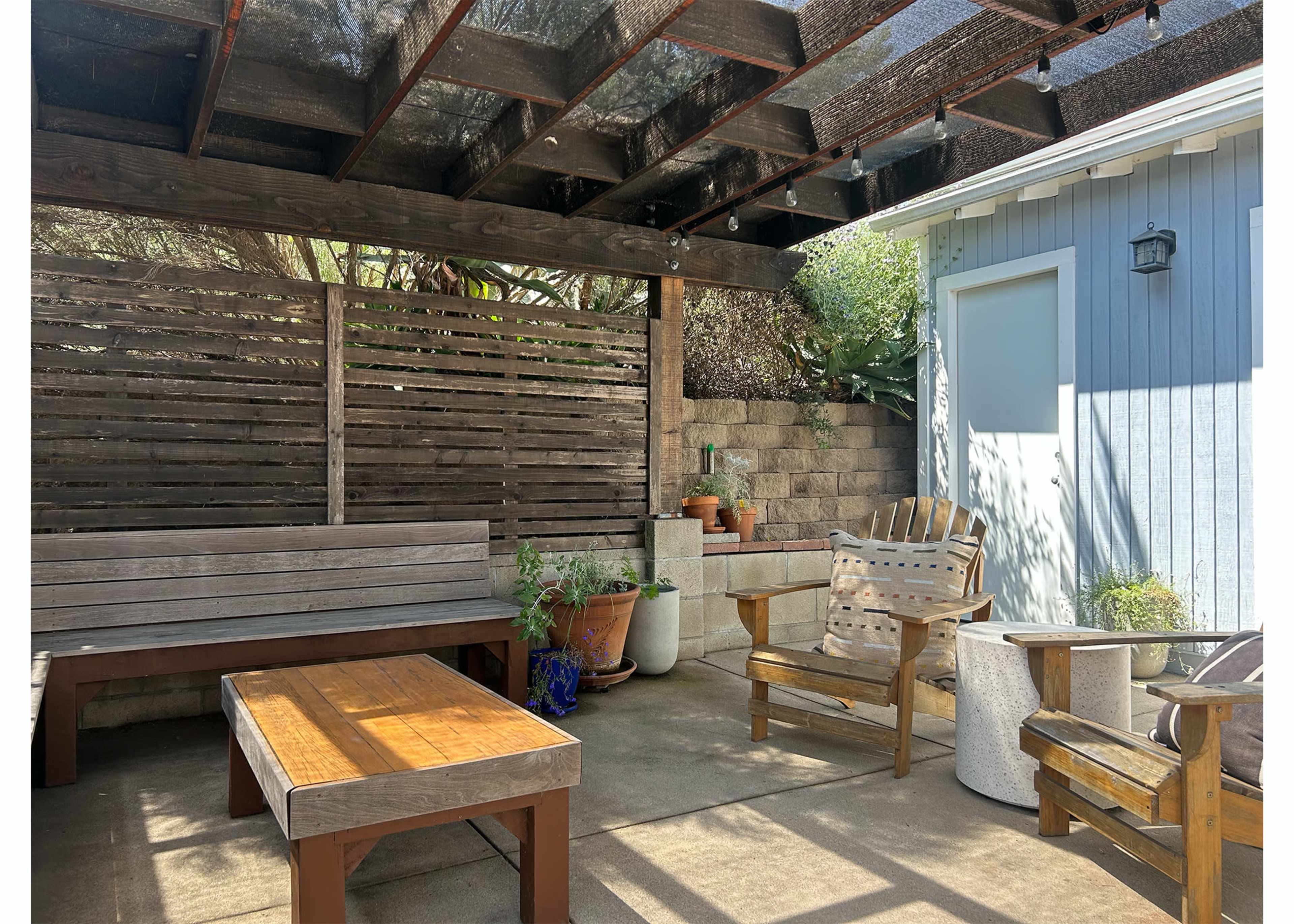The image shows a shaded outdoor seating area with wooden benches, a coffee table, and potted plants, surrounded by a stone wall and a blue house.