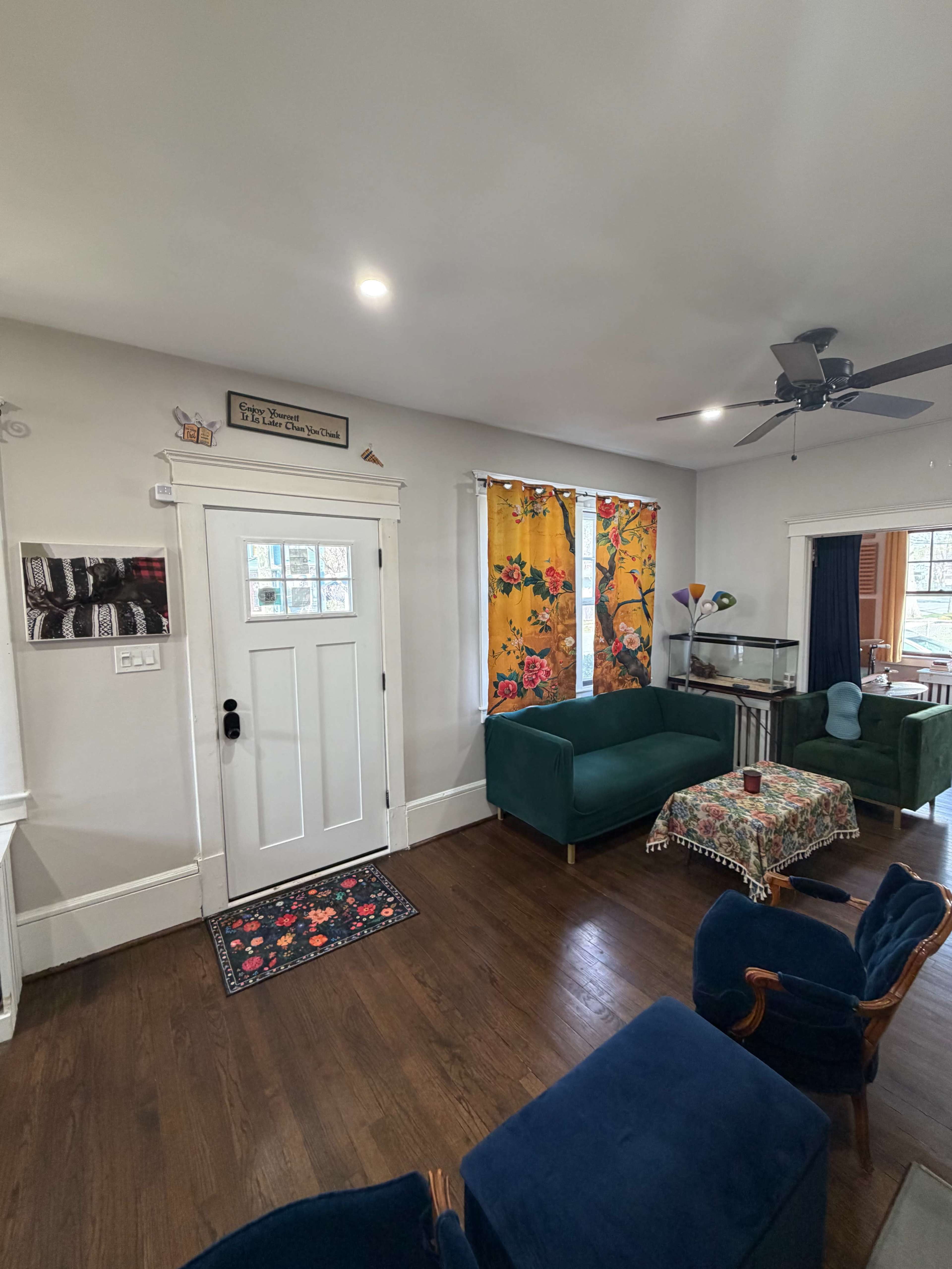 The room features a front door with a colorful rug, a small table with a floral tablecloth, and two green couches set against a wall with decorative curtains.