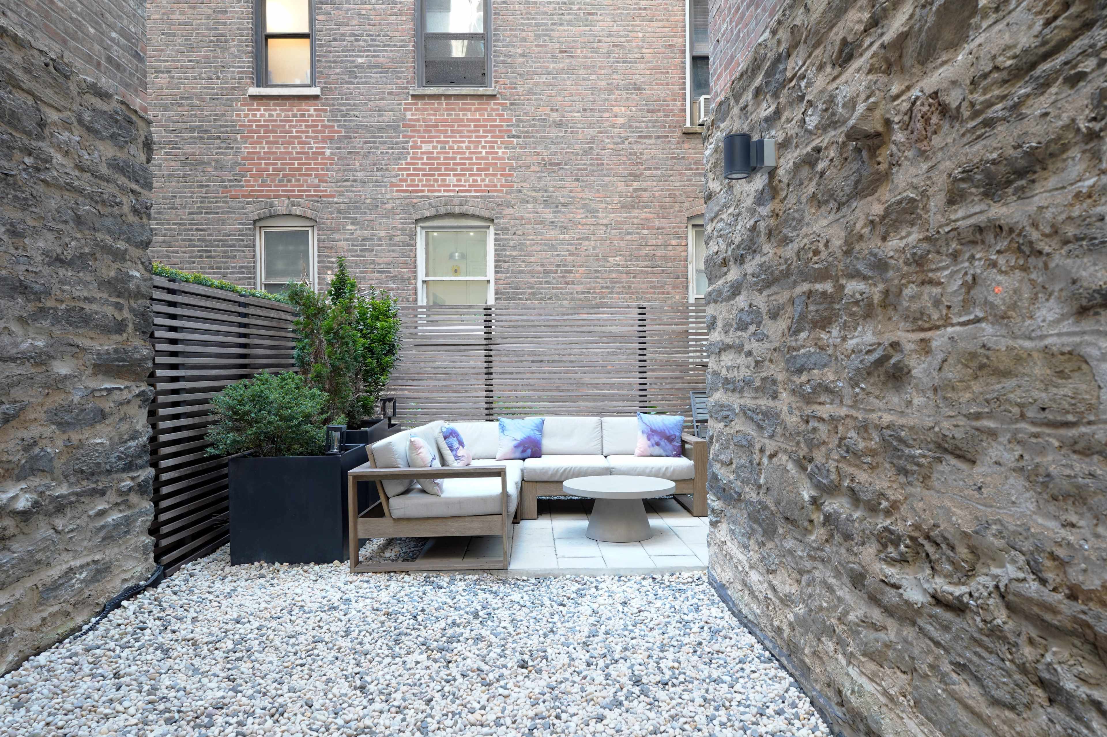 The image shows a narrow outdoor courtyard with stone walls, featuring a seating area with a sofa and a round table surrounded by gravel and planters.