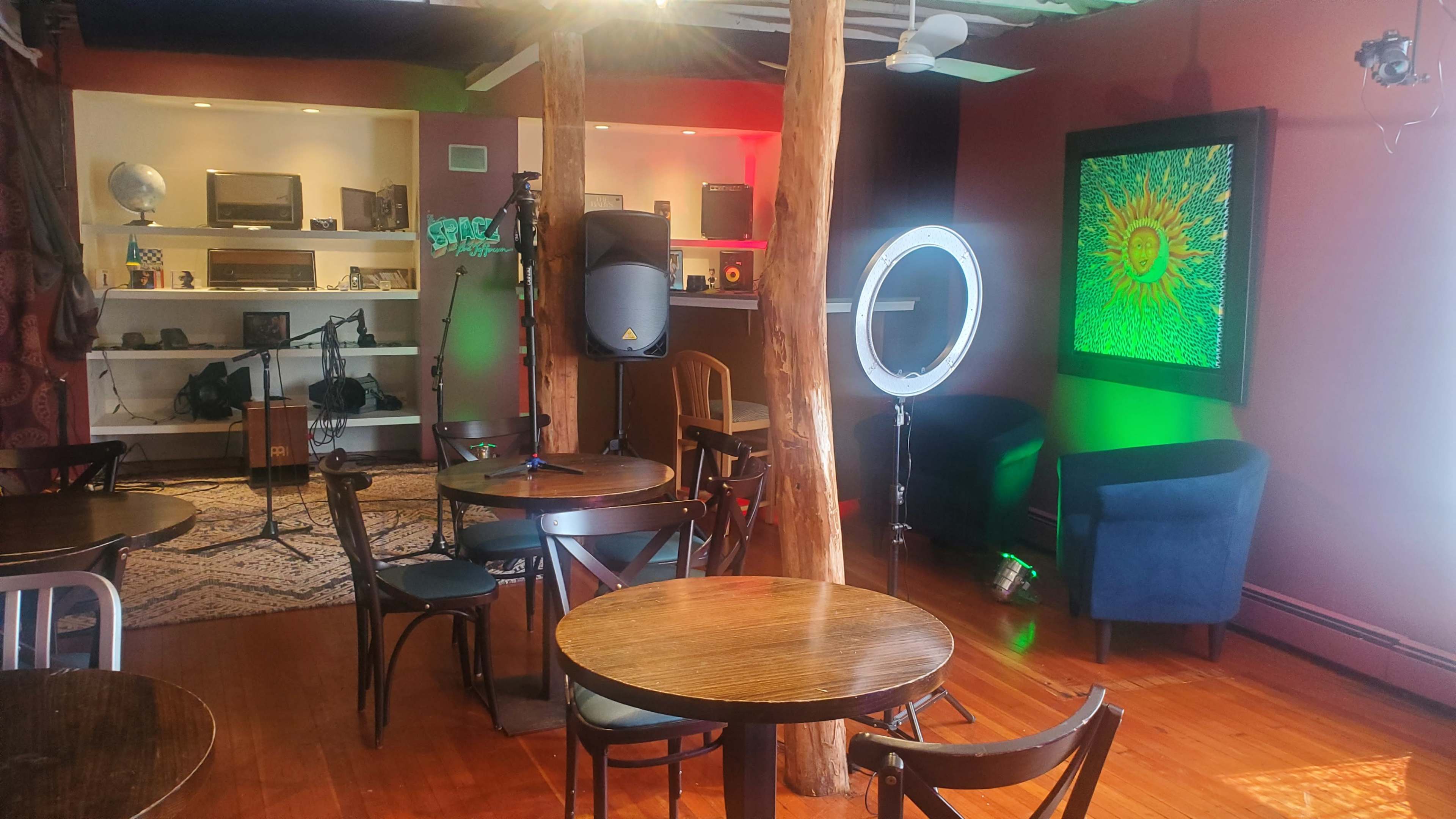 A small performance space with wooden floors, several tables, a microphone setup, and colorful lighting against a backdrop of shelves and art.
