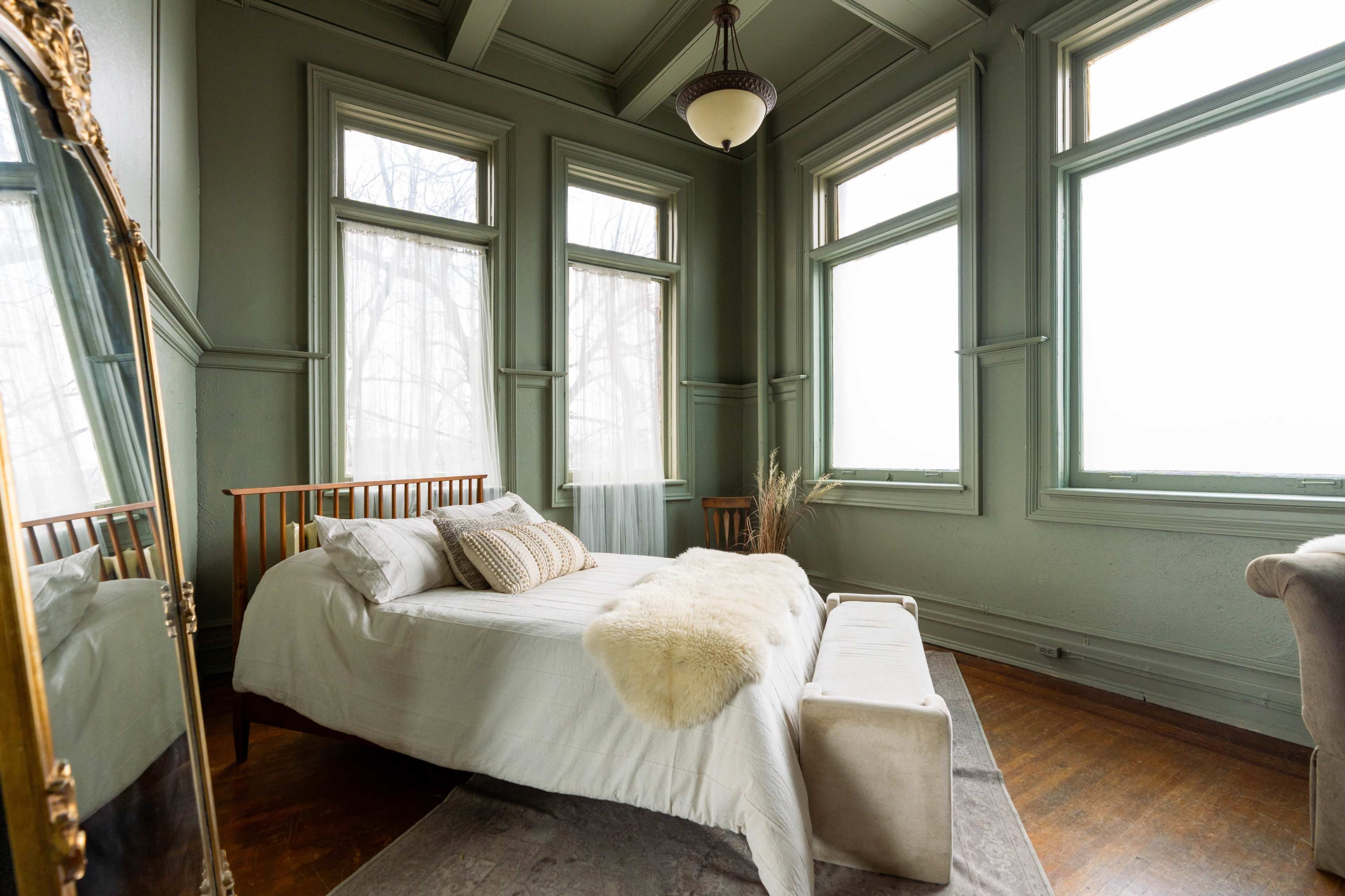 A spacious bedroom features a wooden bed with white bedding, a plush throw, and large windows allowing natural light to filter in.