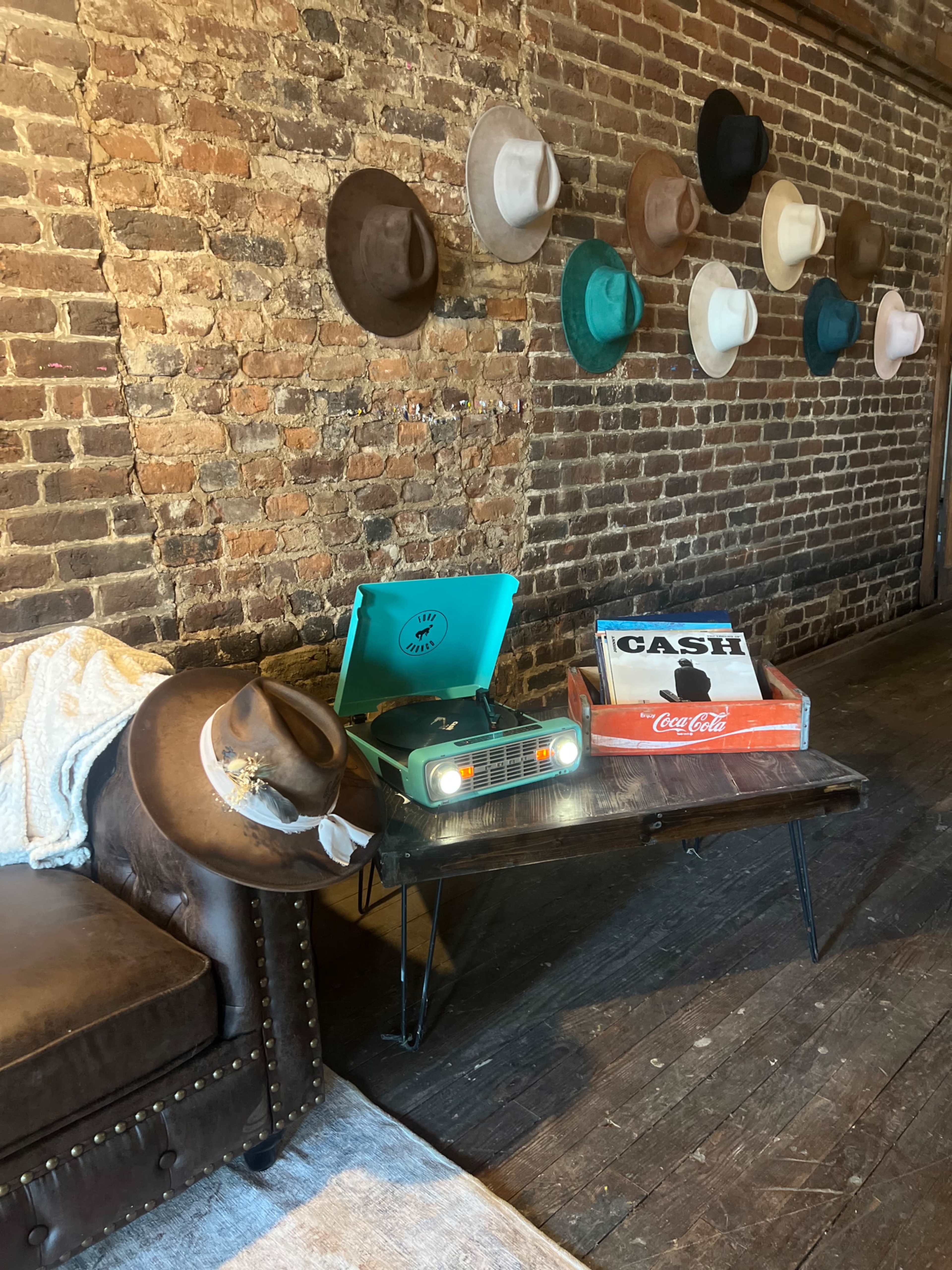 A vintage record player sits on a metal table next to a box labeled "CASH" in a room with an exposed brick wall adorned with various hats.